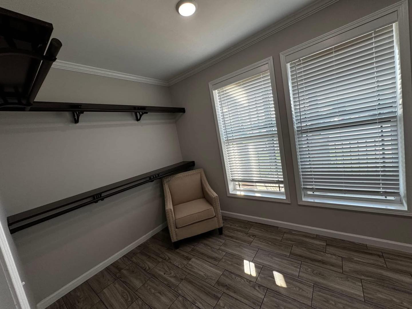 Minimalist room with wooden floor, beige armchair, and dark shelves against a gray wall. Two windows with blinds let in soft natural light.