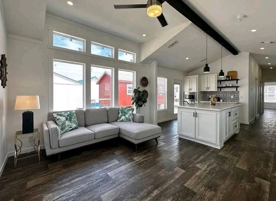 Modern living room and kitchen with a gray L-shaped sofa, tropical cushions, dark wood floor, white kitchen cabinets, large windows, and ceiling fan.