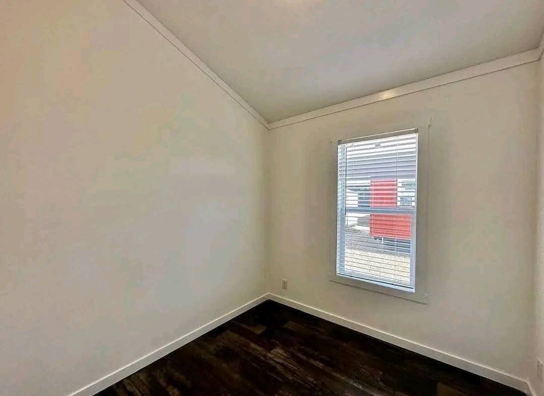 A small, empty room with slanted ceiling, white walls, and dark wooden floor. A window with blinds allows natural light, creating a bright, minimalist atmosphere.