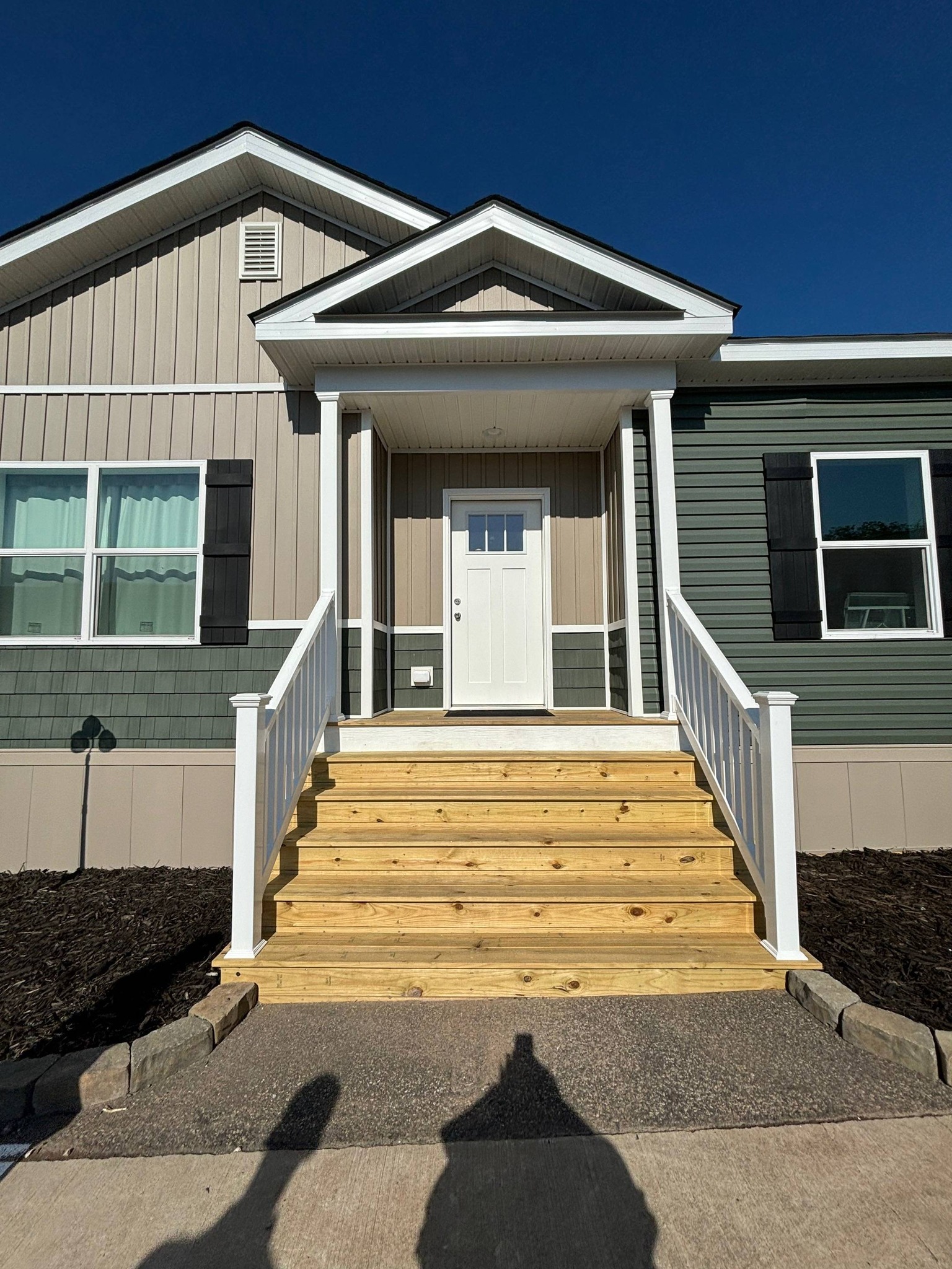 A modern house with teal and tan siding, featuring a white front door and stairs with railings. The clear blue sky creates a bright, welcoming atmosphere.