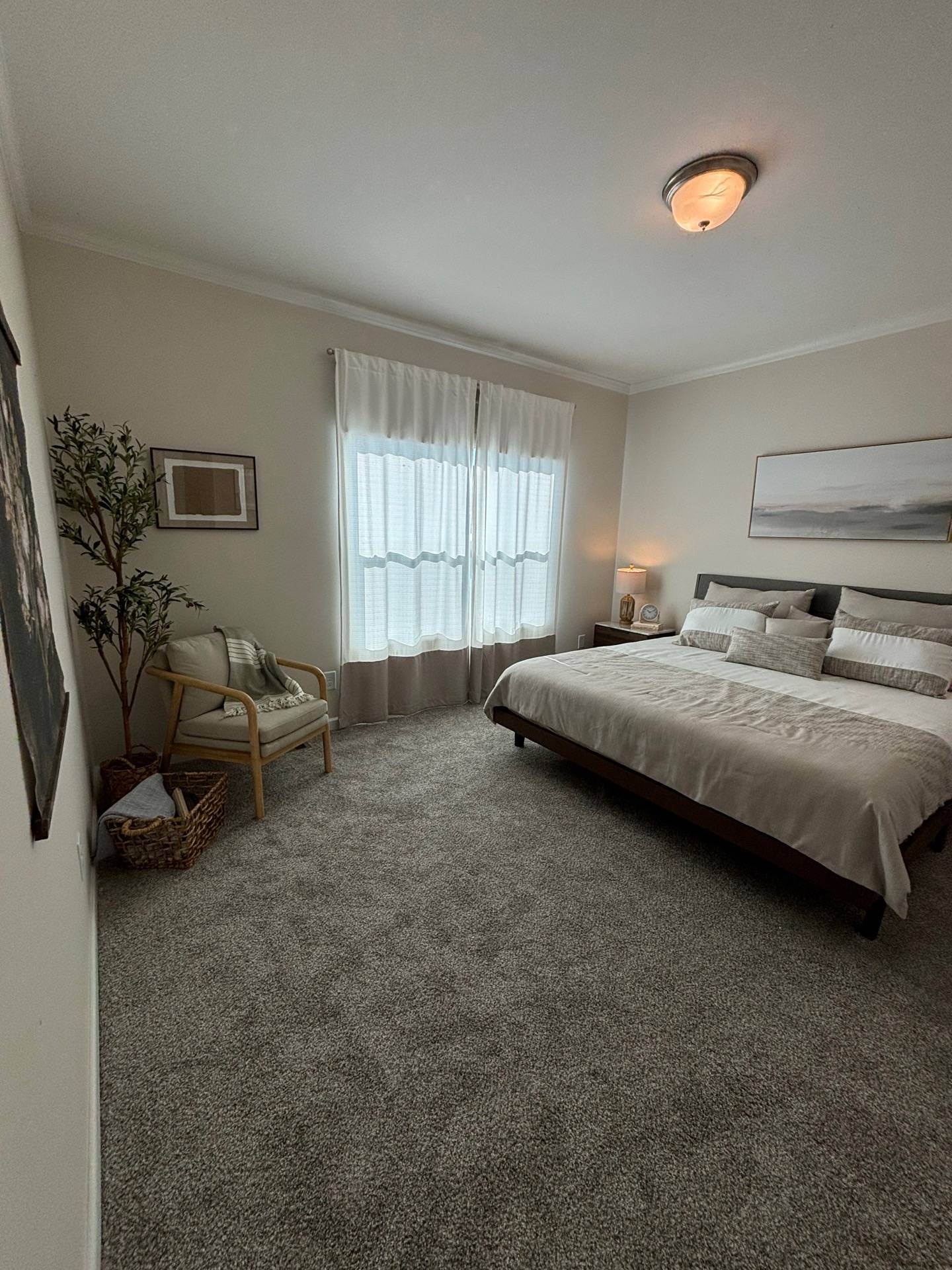 A cozy, minimalist bedroom features a neatly made bed with white linens, soft lighting, a chair with a green throw, sheer curtains, and neutral decor.