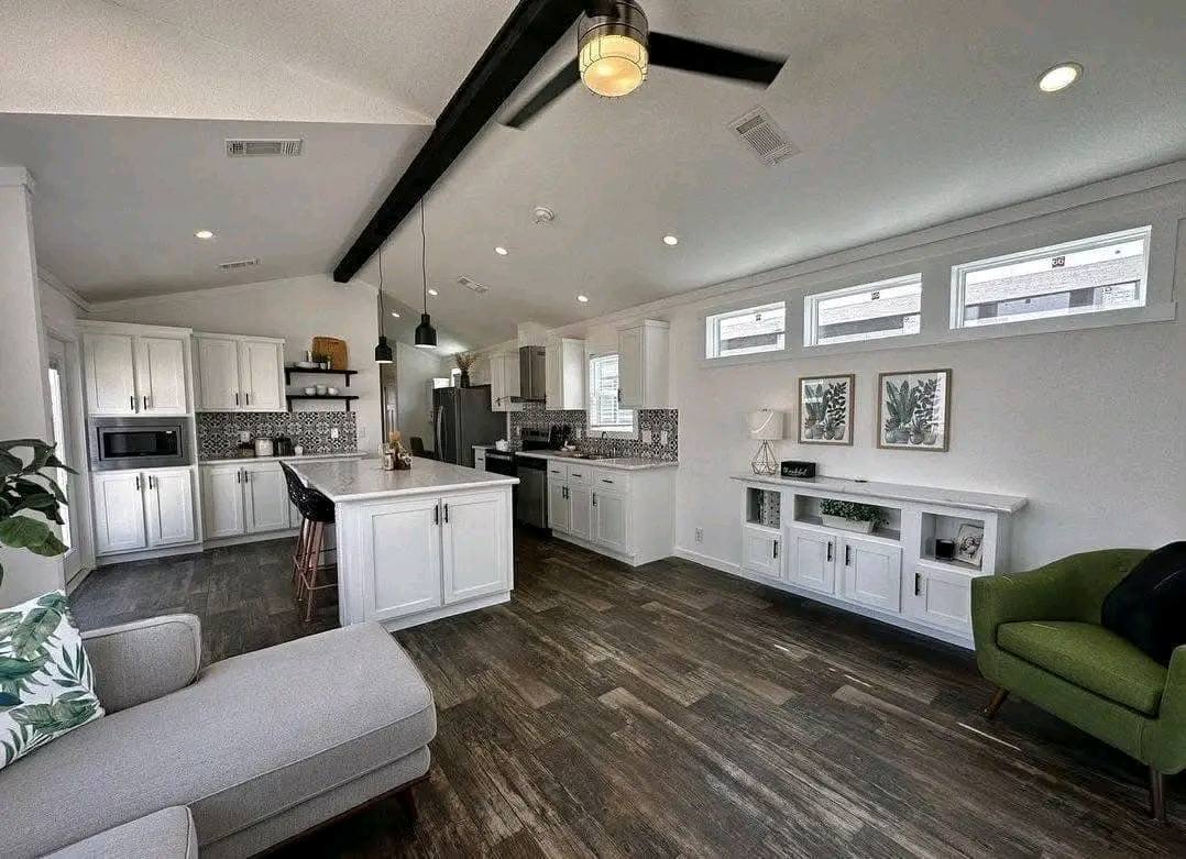 Modern kitchen and living area with dark wood flooring, white cabinets, and black accents. Light streams through high windows, creating a bright and airy feel.