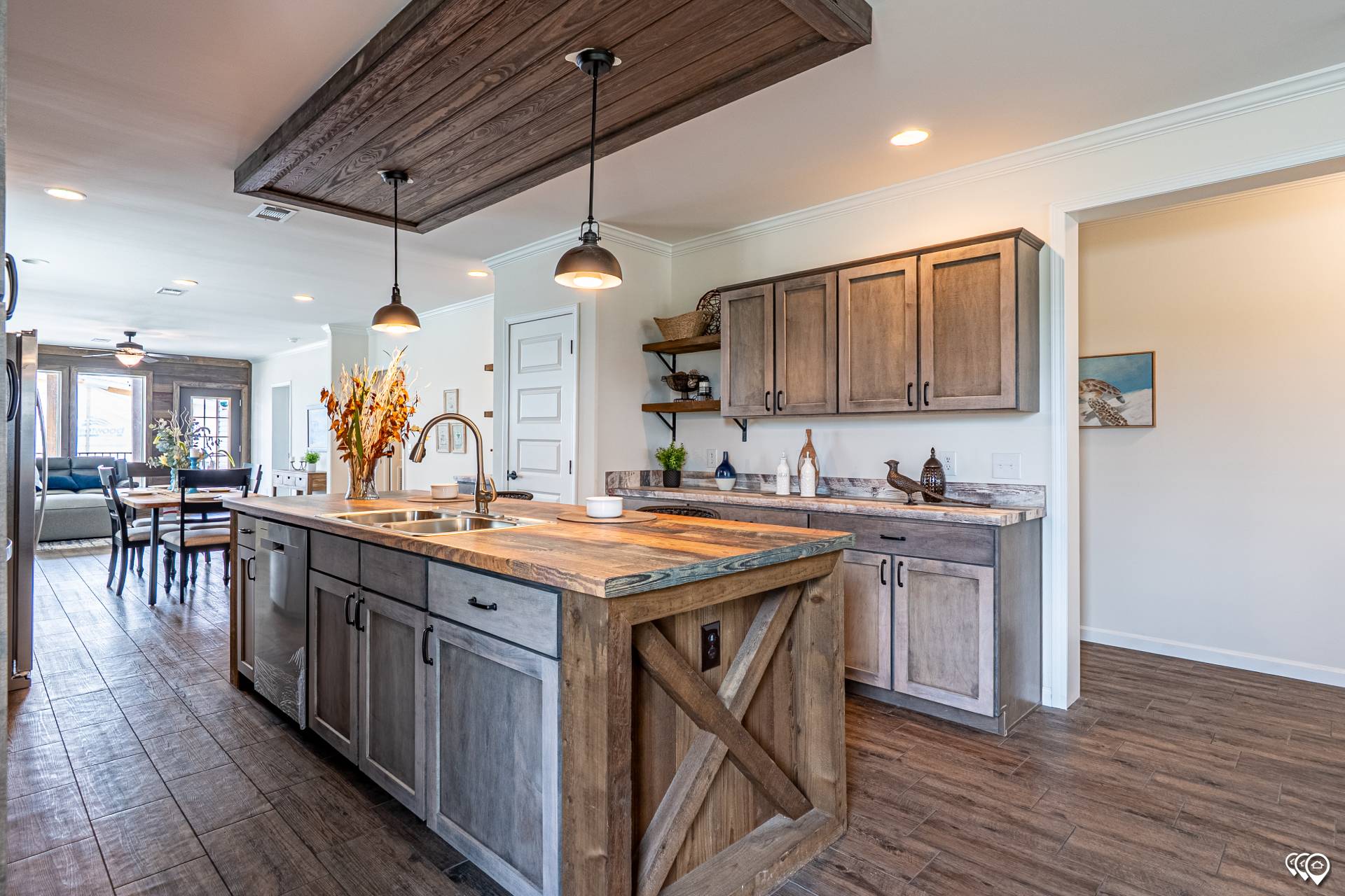 A rustic kitchen features a wooden island with pendant lights, stone countertops, and wood cabinets. Dining area and living space visible in the background.