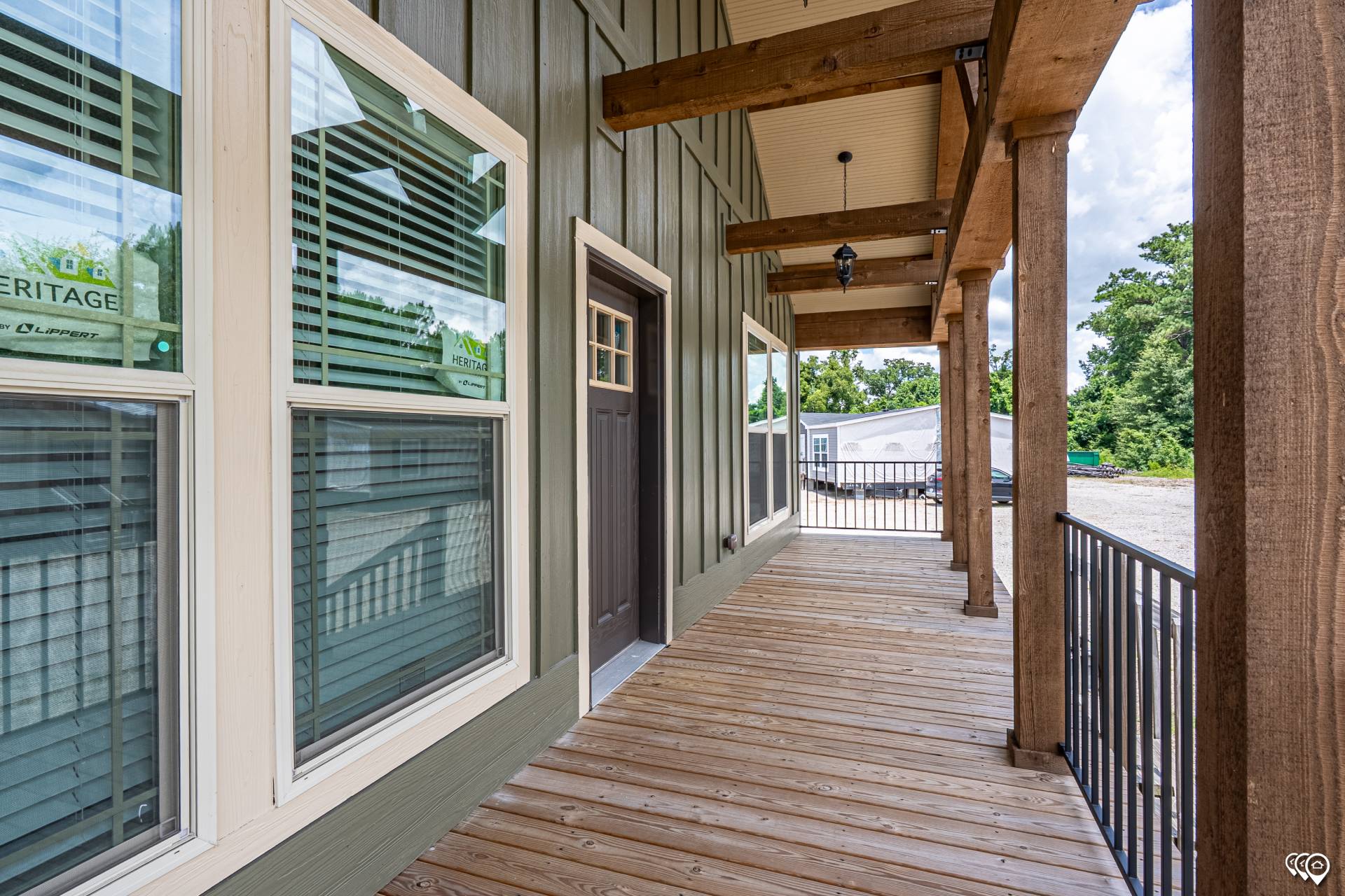 Wooden porch with large windows, dark brown door, and exposed beams. Overlooks a driveway and trees under a partly cloudy sky, creating a rustic feel.
