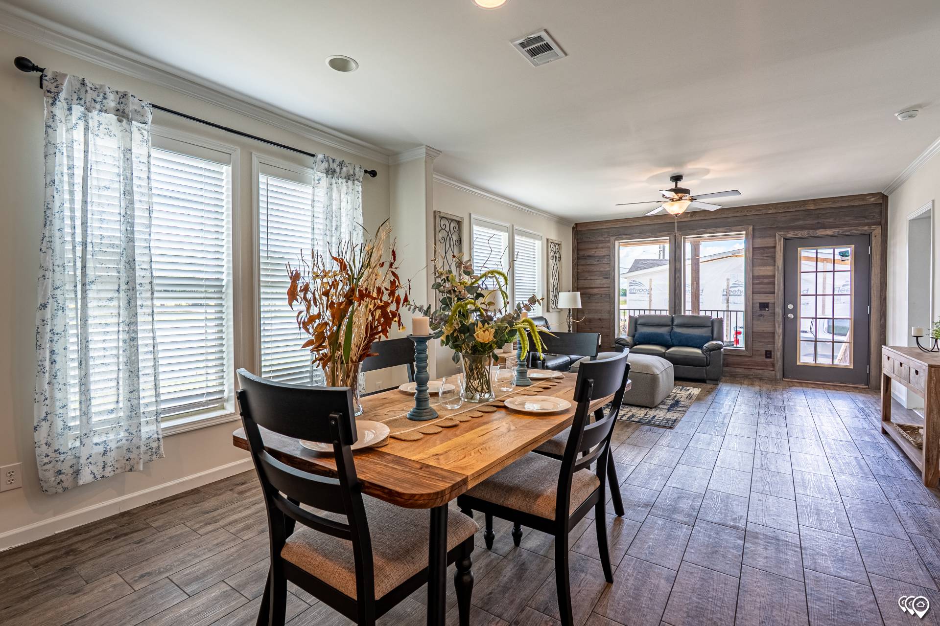 Open-concept living and dining room with wooden table, chairs, and rustic decor. Natural light streams through large windows, creating a cozy ambiance.