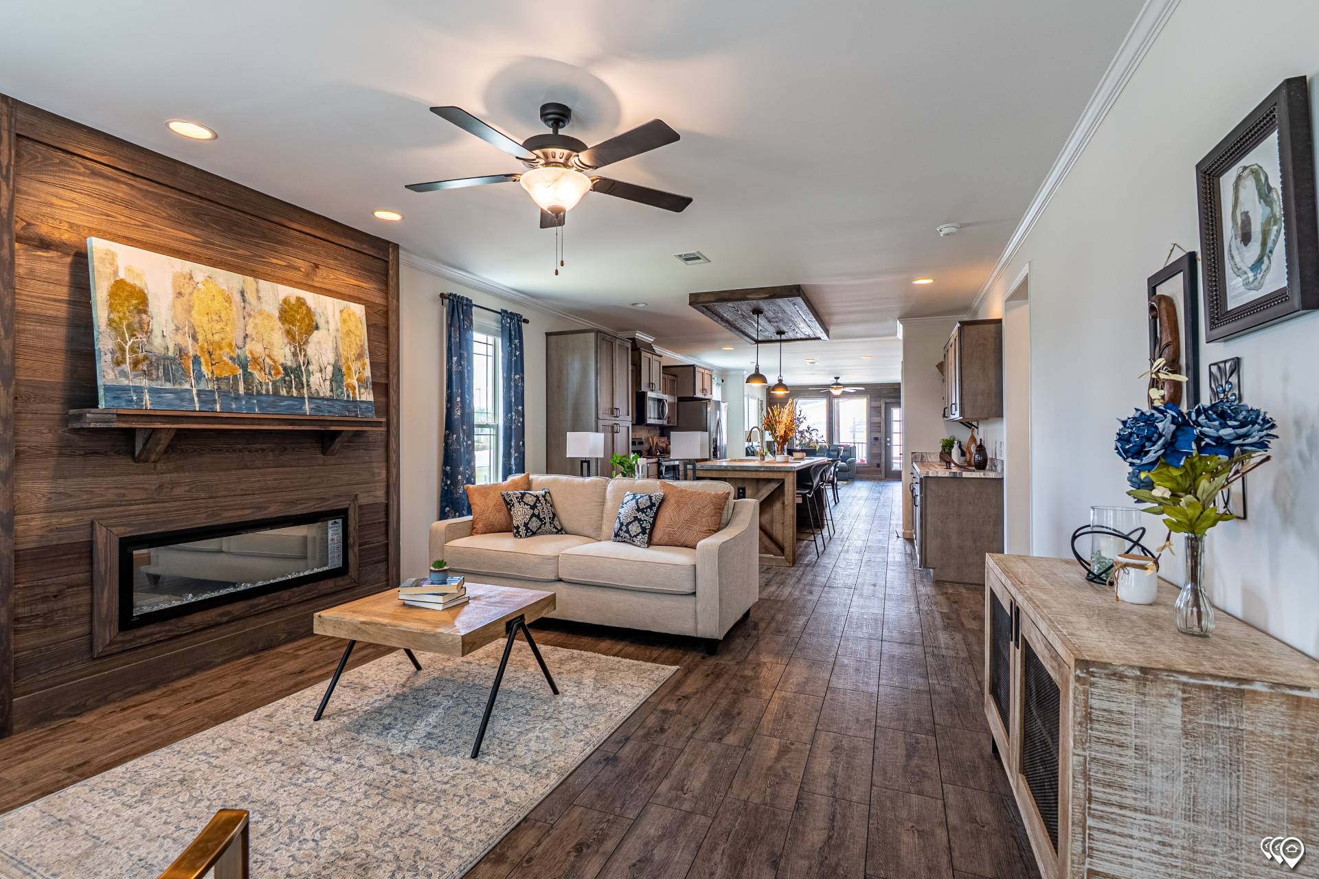 Modern living room with wooden floors, beige sofa, and a coffee table. A ceiling fan, fireplace, and artwork create a cozy feel. Open-concept kitchen in the background.