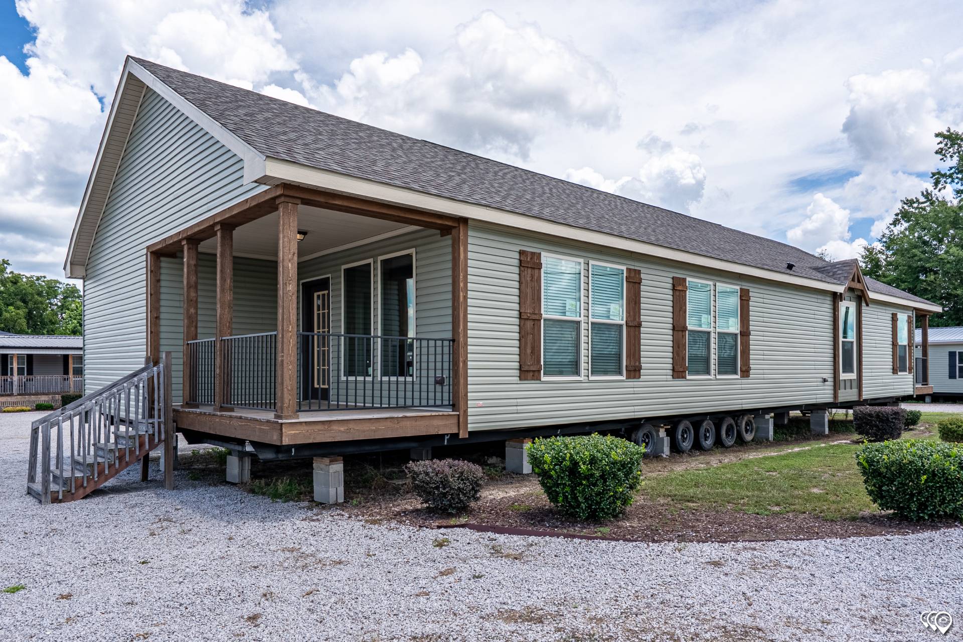 A beige mobile home on wheels with a wooden porch and railing, against a cloudy sky. Gravel landscaping surrounds the area, creating a serene, rural feel.