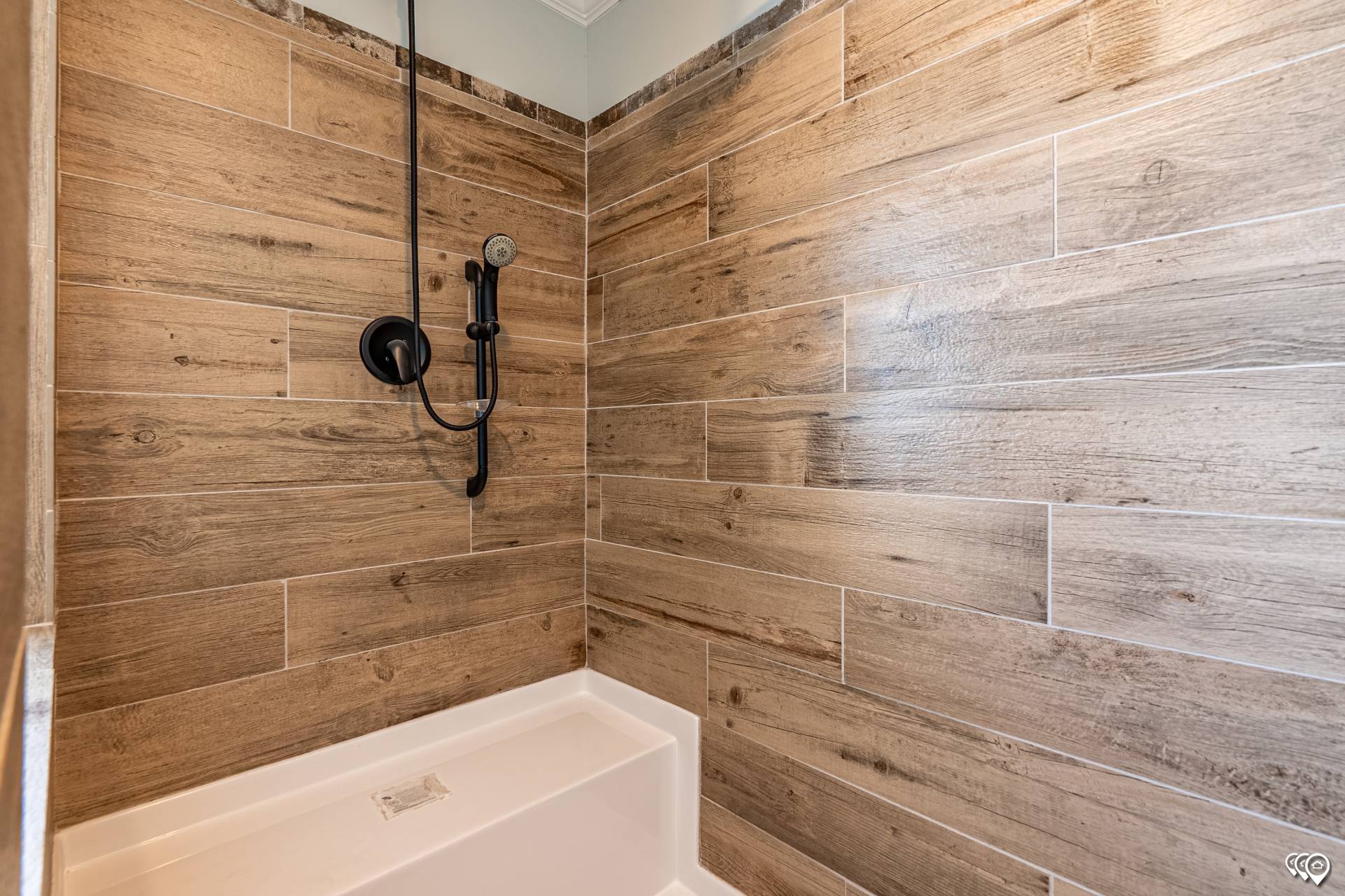A shower interior with wood-textured tiles on the walls, featuring a matte black showerhead and controls. The tone is modern and rustic.