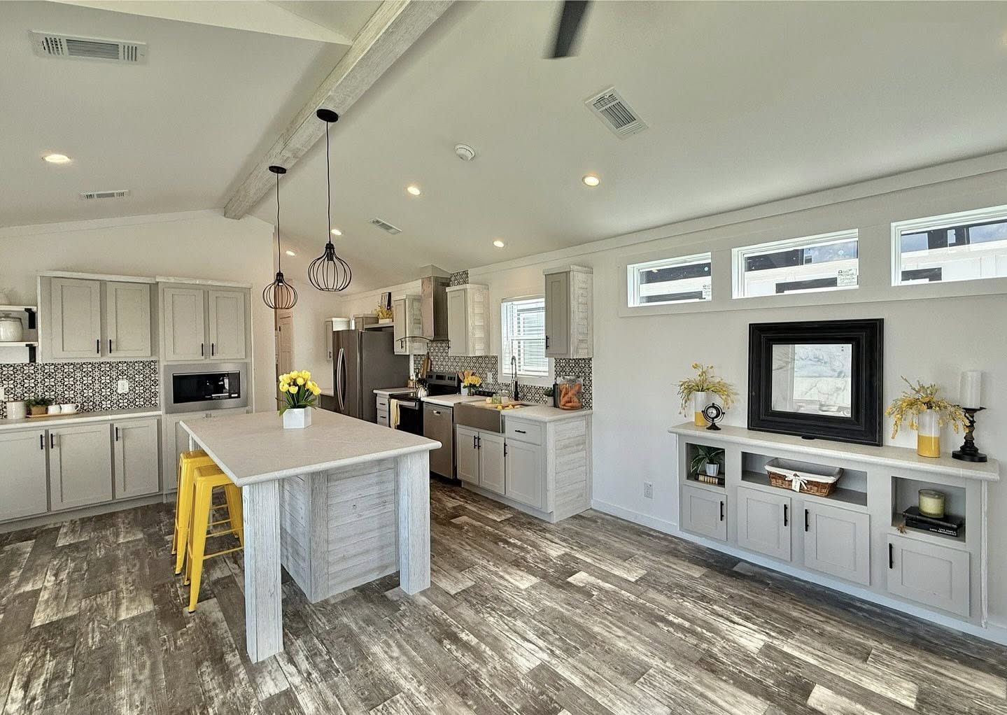 Spacious modern kitchen with light gray cabinets, rustic wood floor, and island with yellow stools. Pendant lights add warmth and style. Bright and inviting.