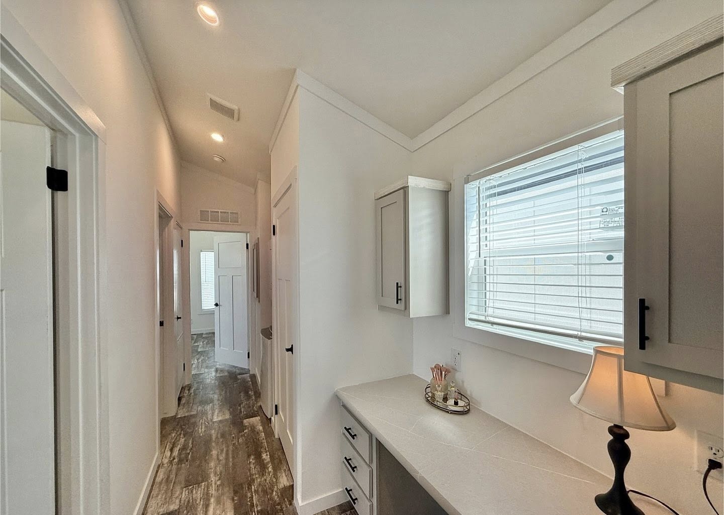 Bright hallway with white walls and wood flooring, featuring a side table with a lamp and tray under a window with blinds. Cozy and inviting atmosphere.