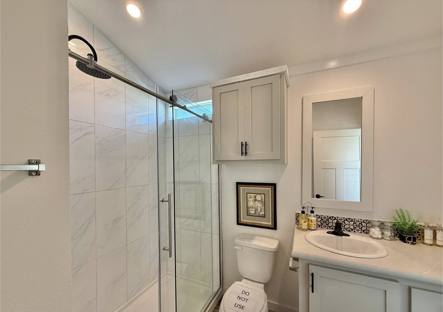 Modern bathroom with a glass-door shower, marble tiles, and recessed lighting. Features a vanity with a mirror and decorated with plants and toiletries.