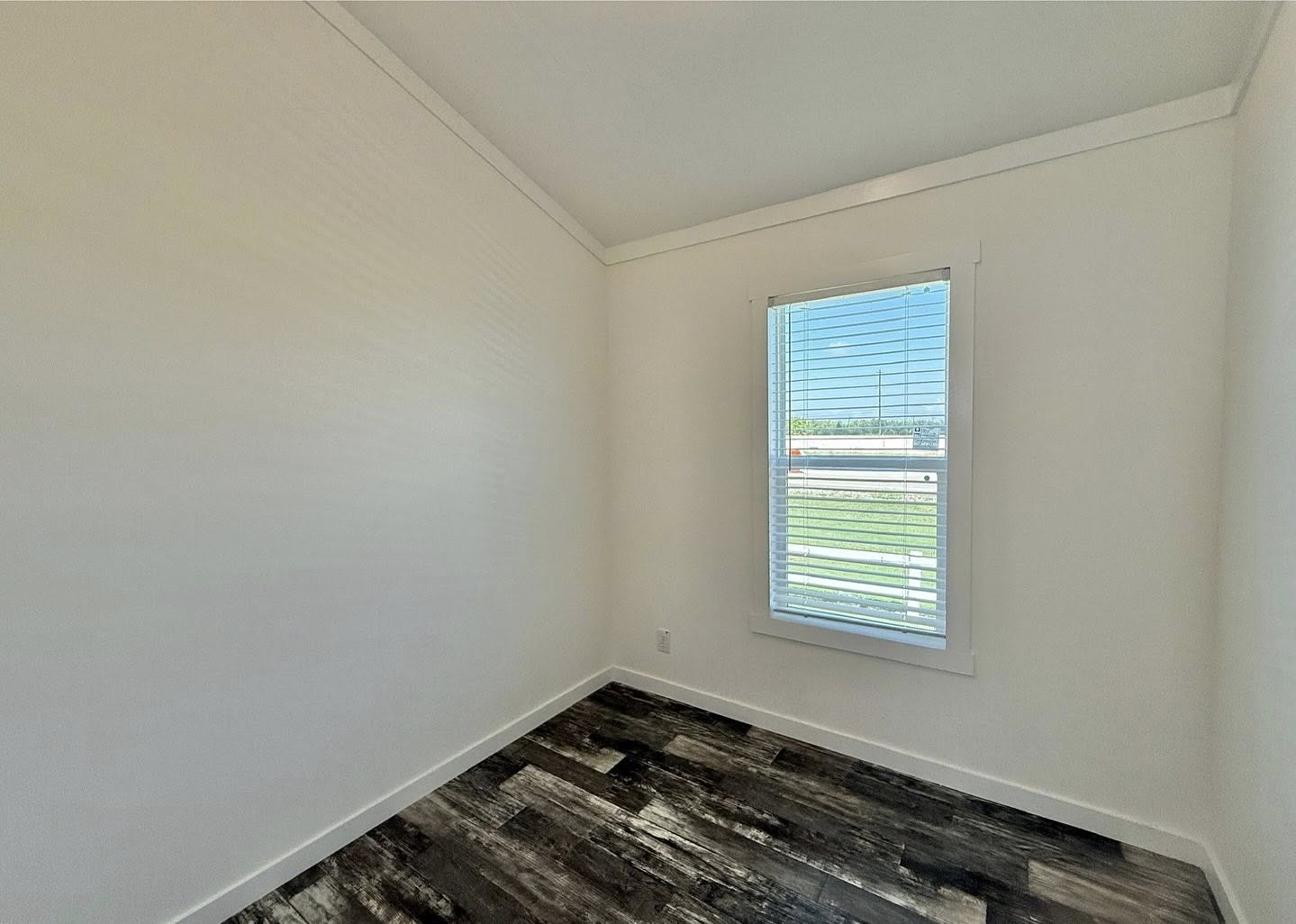 Small, empty room with white walls and a window showing a sunny, outdoor scene. Dark wooden floor adds contrast. Bright and minimalist ambiance.