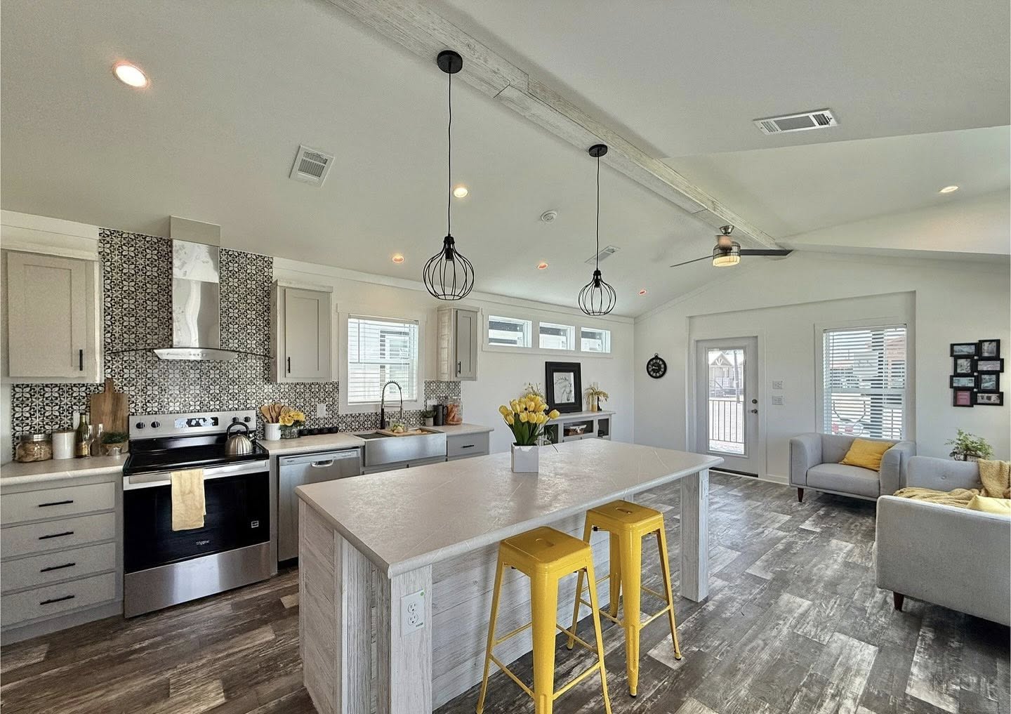 Modern kitchen with a large island, two yellow stools, and hanging pendant lights. Gray cabinets, patterned backsplash, and cozy living area. Bright and inviting.