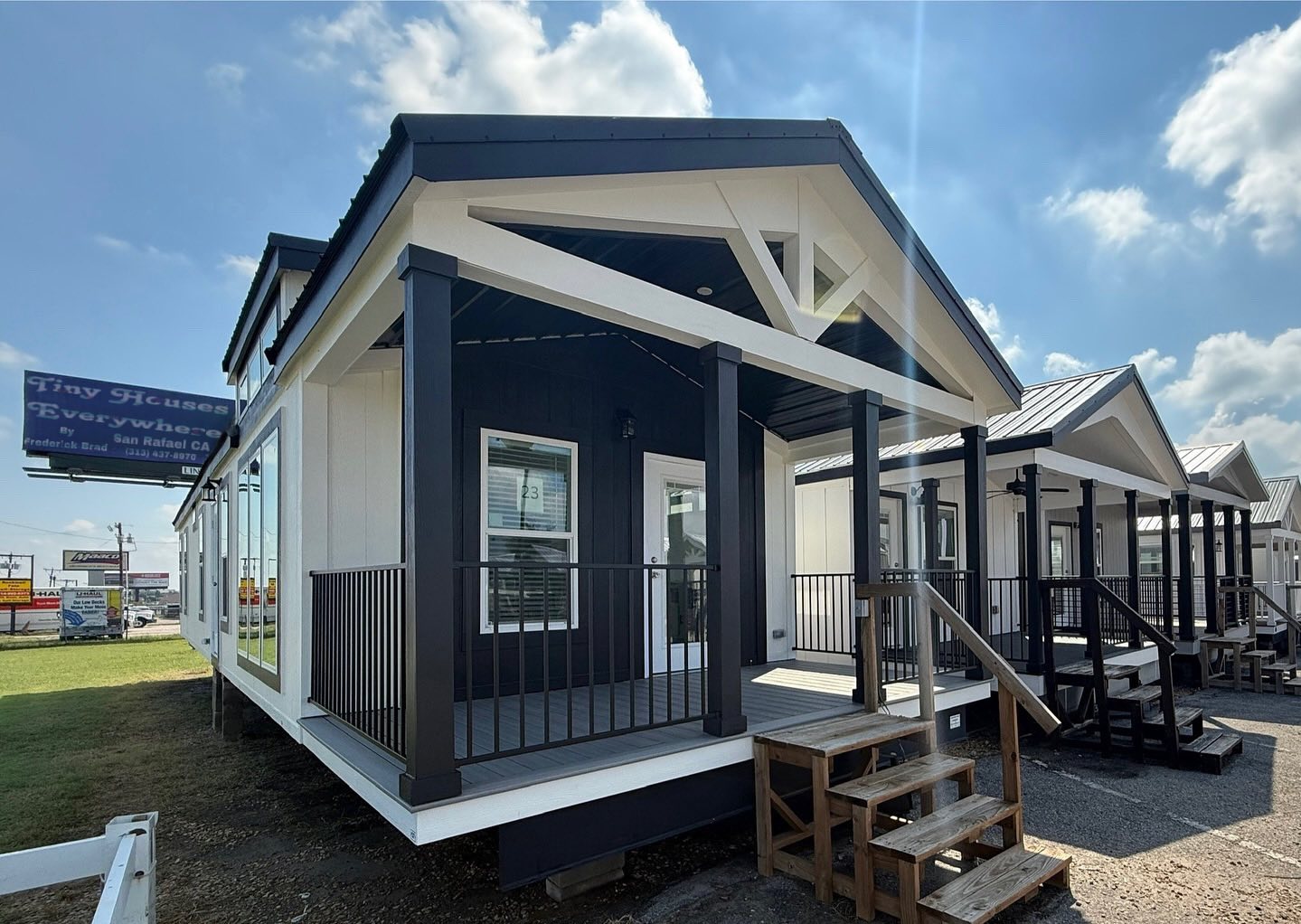 Modern tiny house with a black and white exterior and a small porch. Multiple homes line up under a bright blue sky, creating a compact community feel.