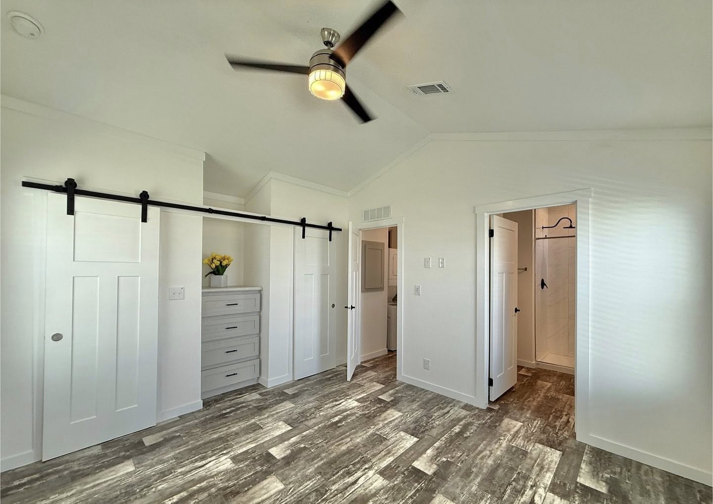 A bright, modern room with a wooden floor, ceiling fan, and white walls. Features include sliding barn doors, a dresser with flowers, and a doorway leading to a bathroom.