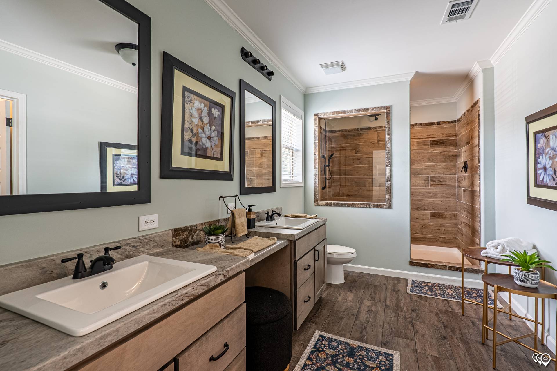 Modern bathroom with dual sinks, black fixtures, and floral artwork. Features a spacious wooden-accented shower, toilet, and decorative plants.