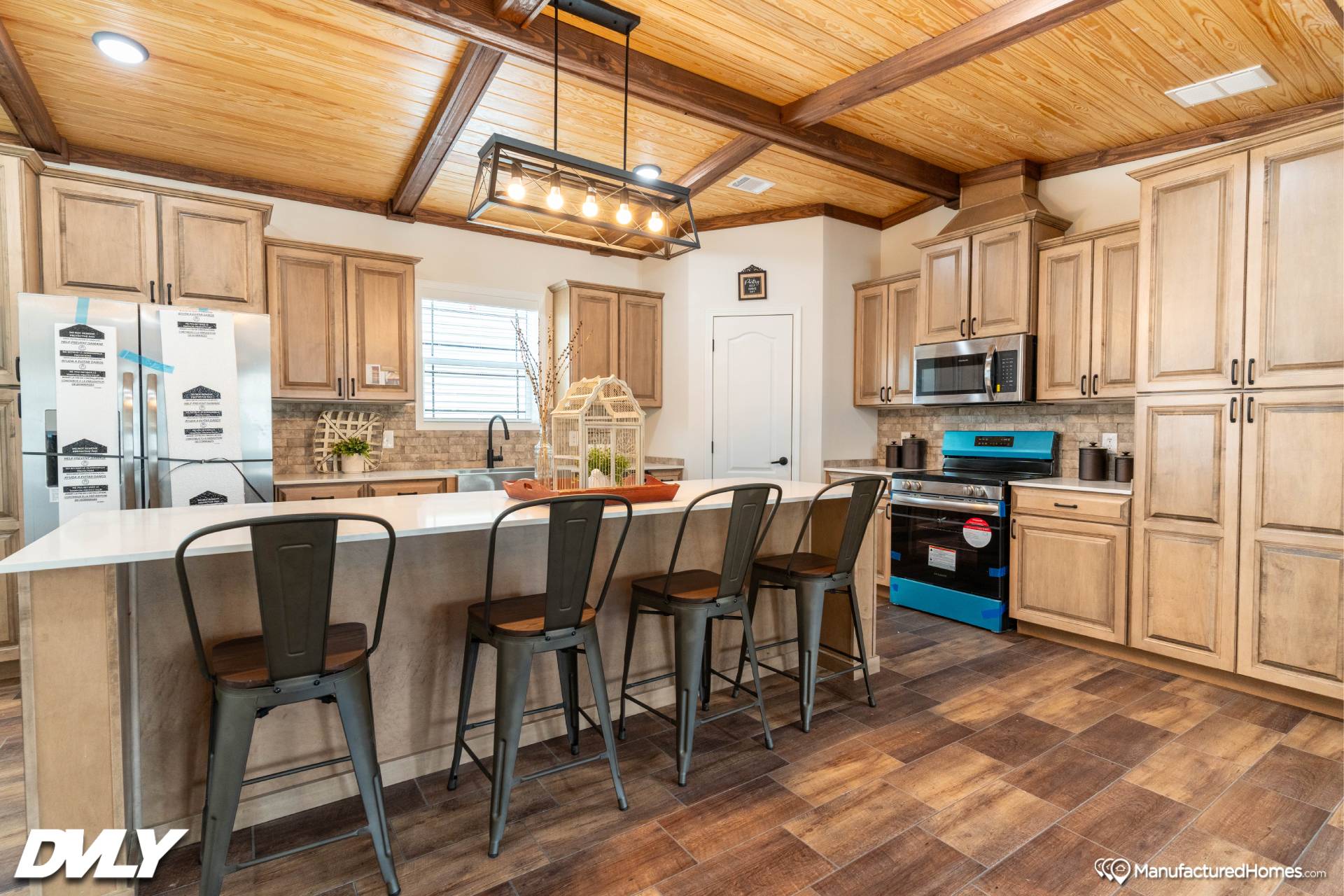 Rustic kitchen with wooden cabinets, a large island with four metal stools, and modern appliances. Warm lighting and wood-paneled ceiling create a cozy feel.