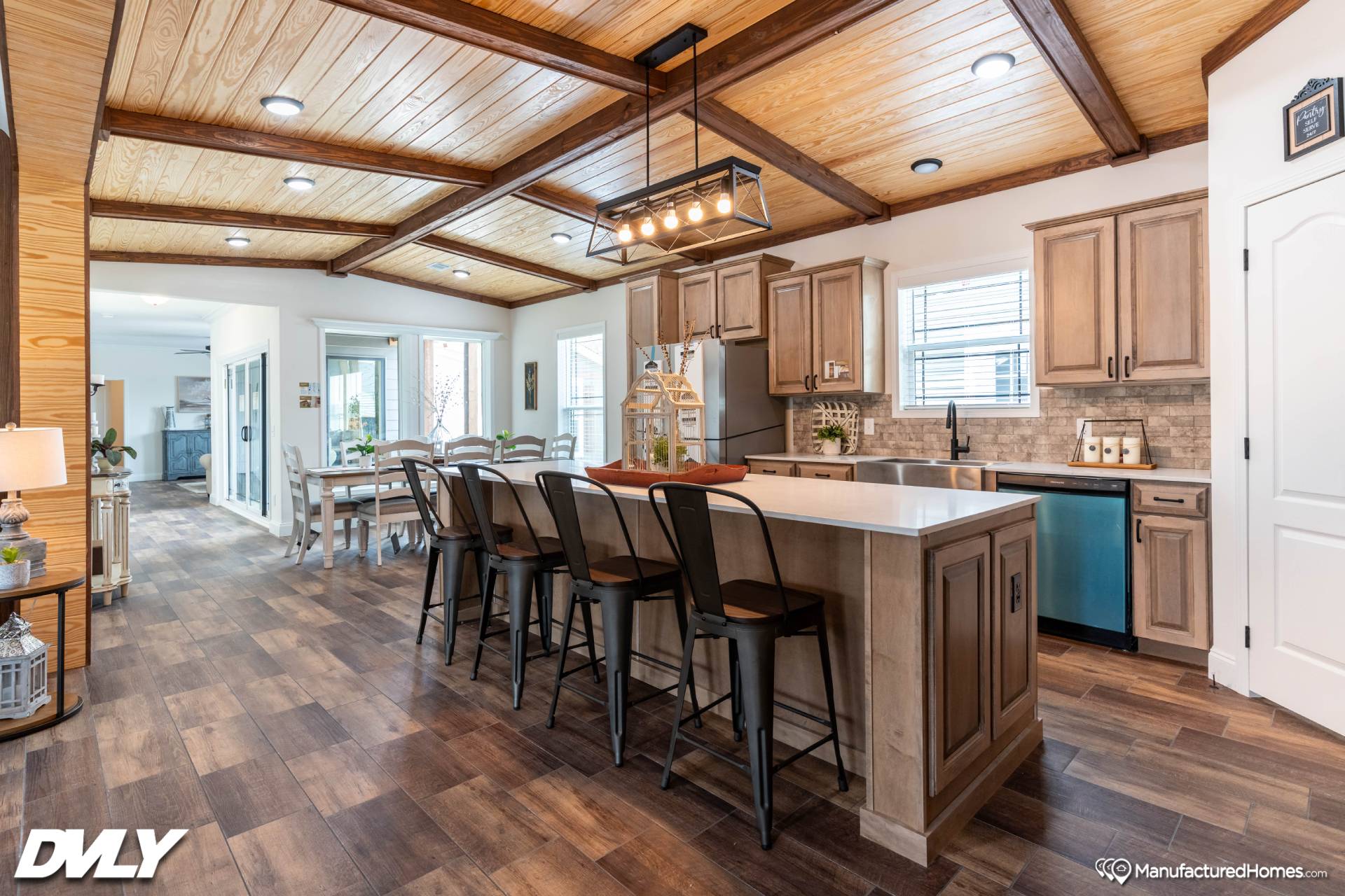 Spacious kitchen with wooden cabinetry, center island, and industrial bar stools. Bright light streams through windows, creating a warm, inviting atmosphere.