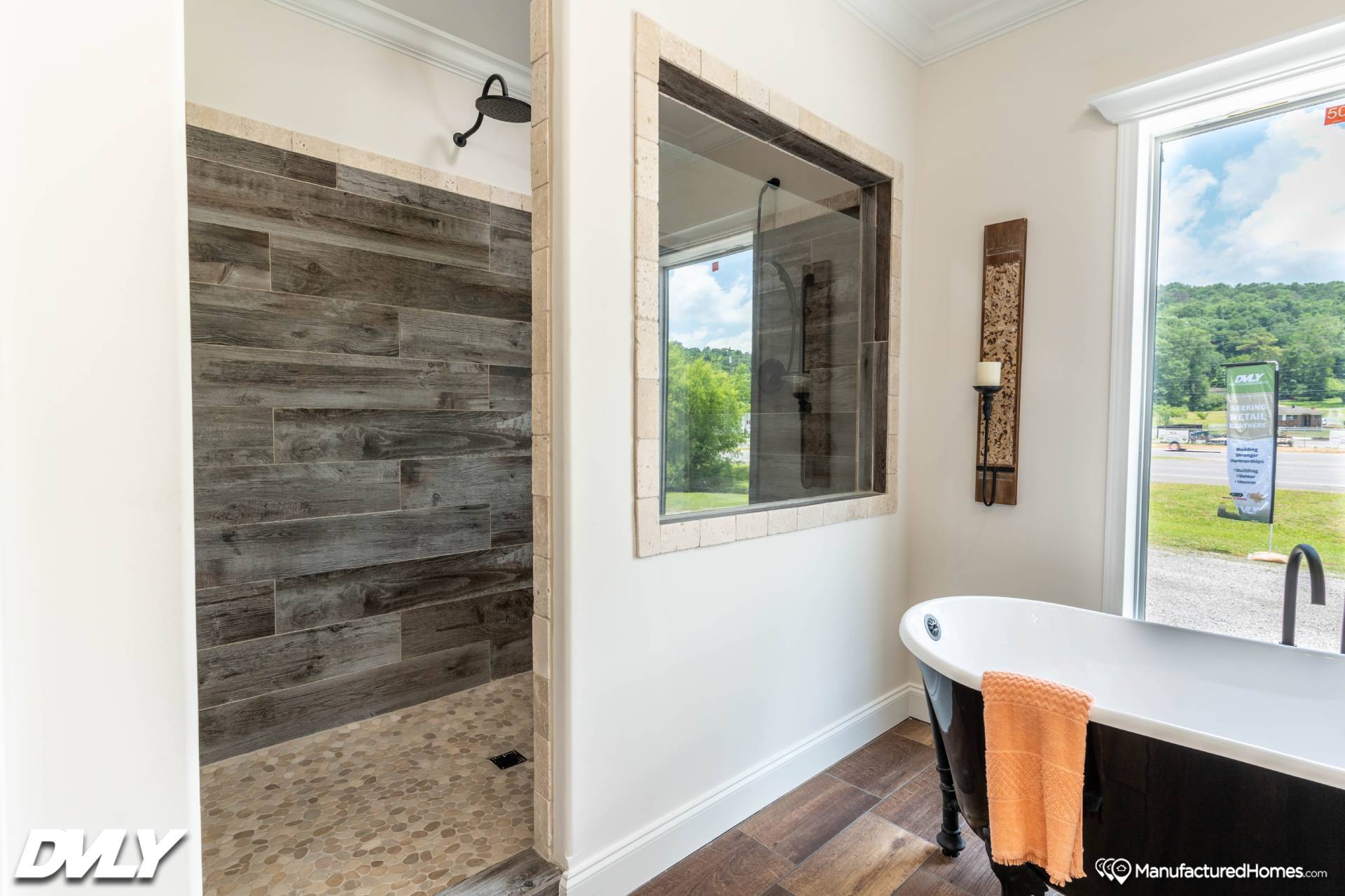 Modern bathroom with a wood-paneled walk-in shower and pebble floor. A freestanding tub with an orange towel is beside a large window, creating an airy feel.