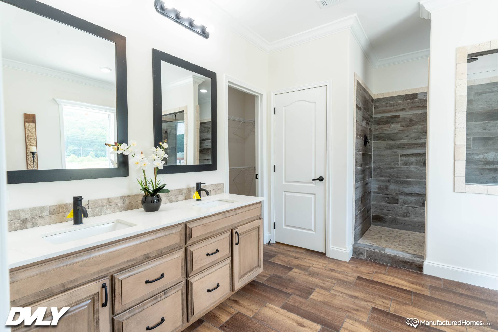 Modern bathroom with light wood cabinetry, dual sinks, large mirrors, and a stone-tiled shower. Features elegant wood flooring and orchid decor.