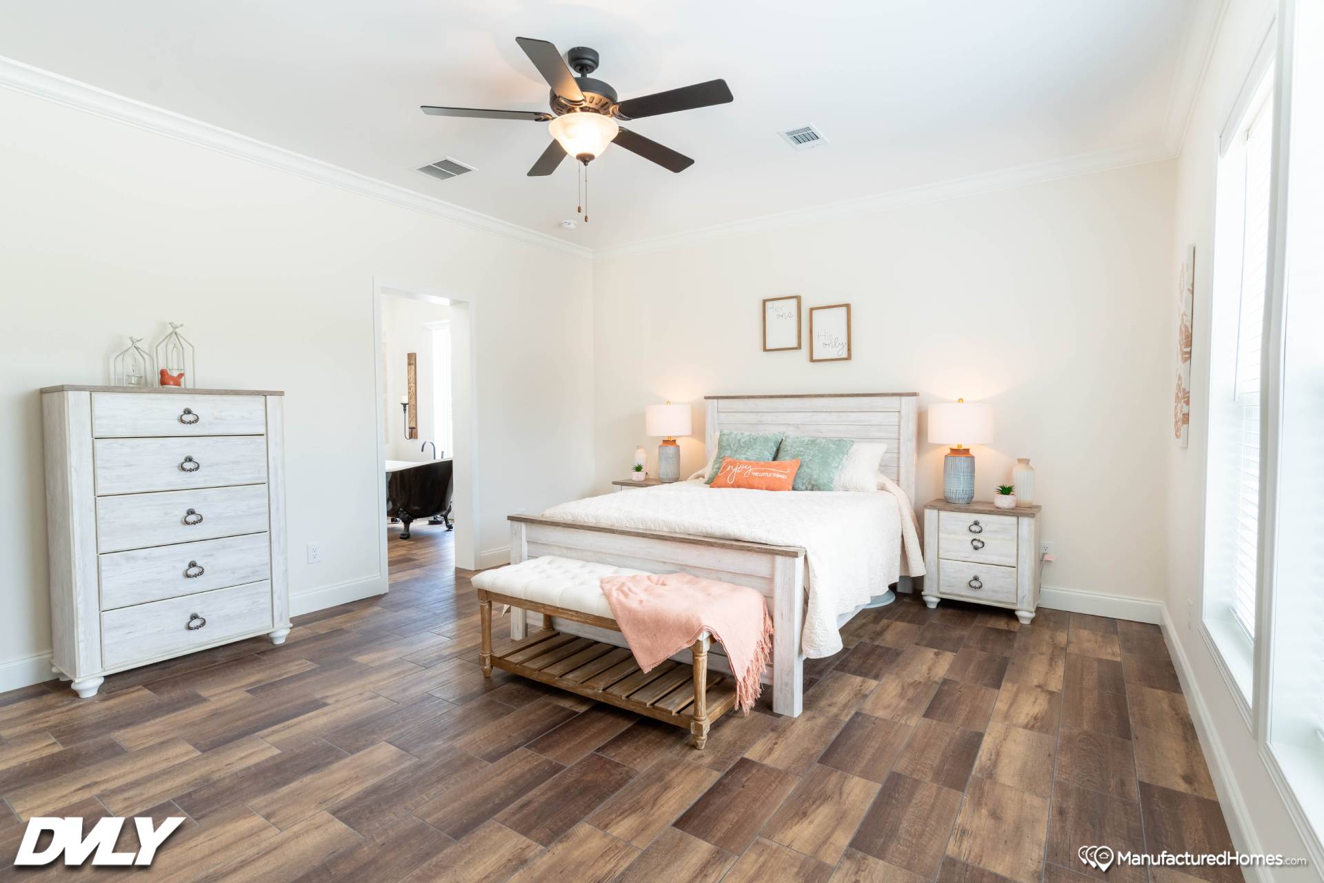 Bright bedroom with wood flooring, featuring a bed with pastel pillows and an orange throw, flanked by nightstands. A dresser stands to the side. Cozy and inviting.