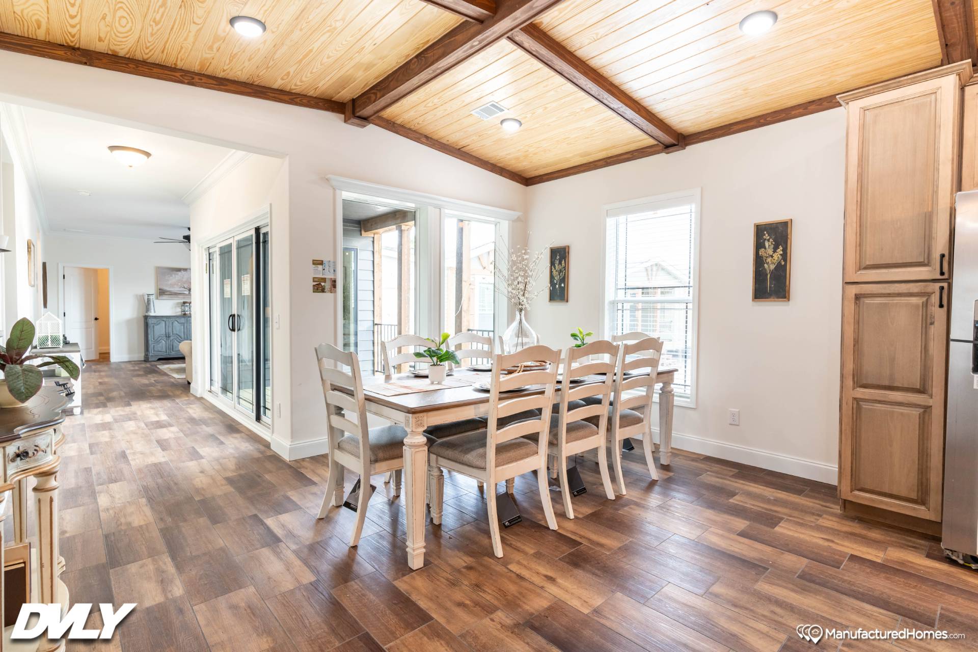 Bright dining area with a wooden table set for six, under a wood-beamed ceiling. Windows bring in natural light; adjacent cozy hallway leads to the kitchen.