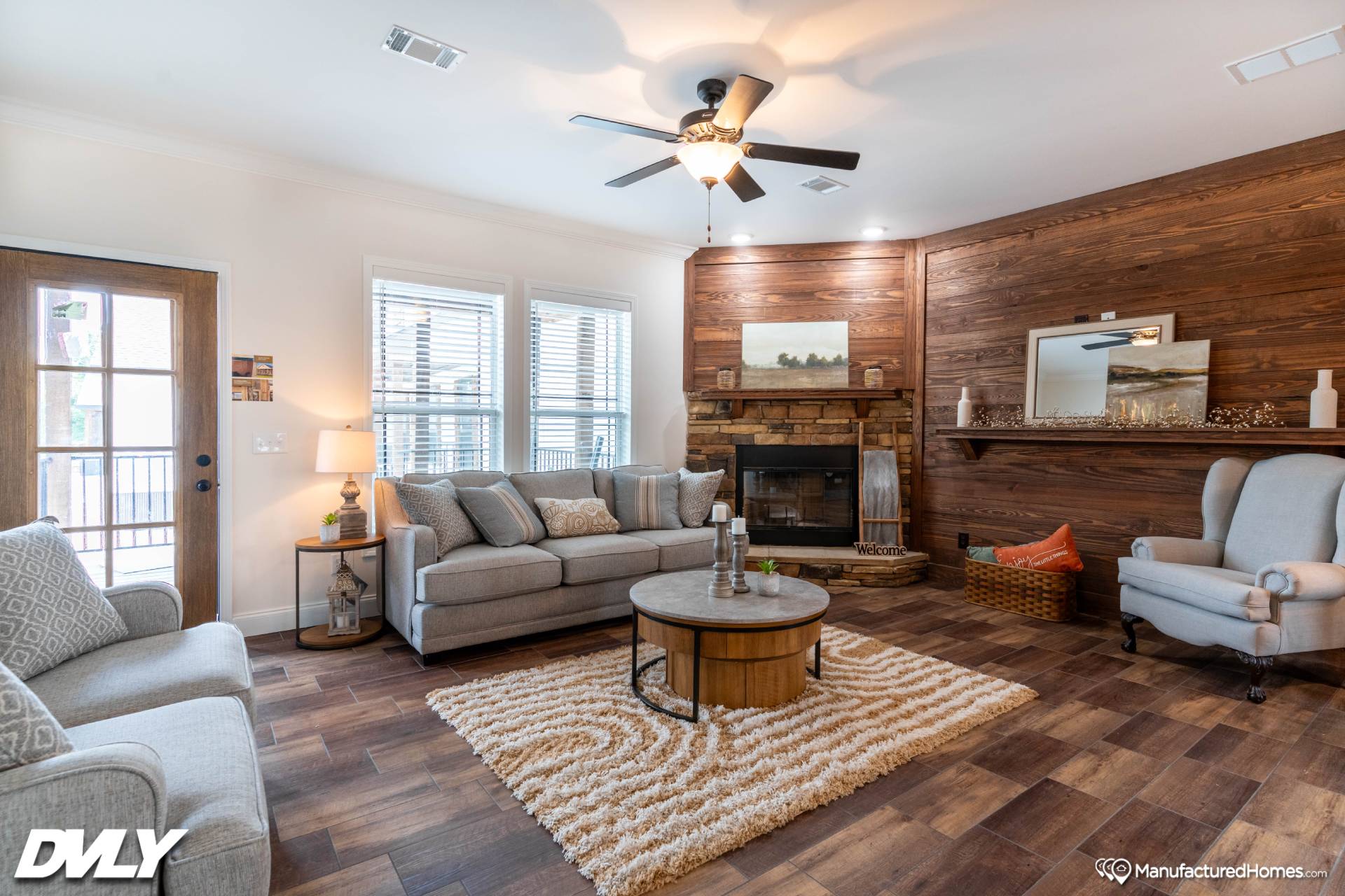 Cozy living room with a gray sofa, armchairs, and wood-paneled walls. A fireplace, circular table, and plush rug enhance the warm ambiance.