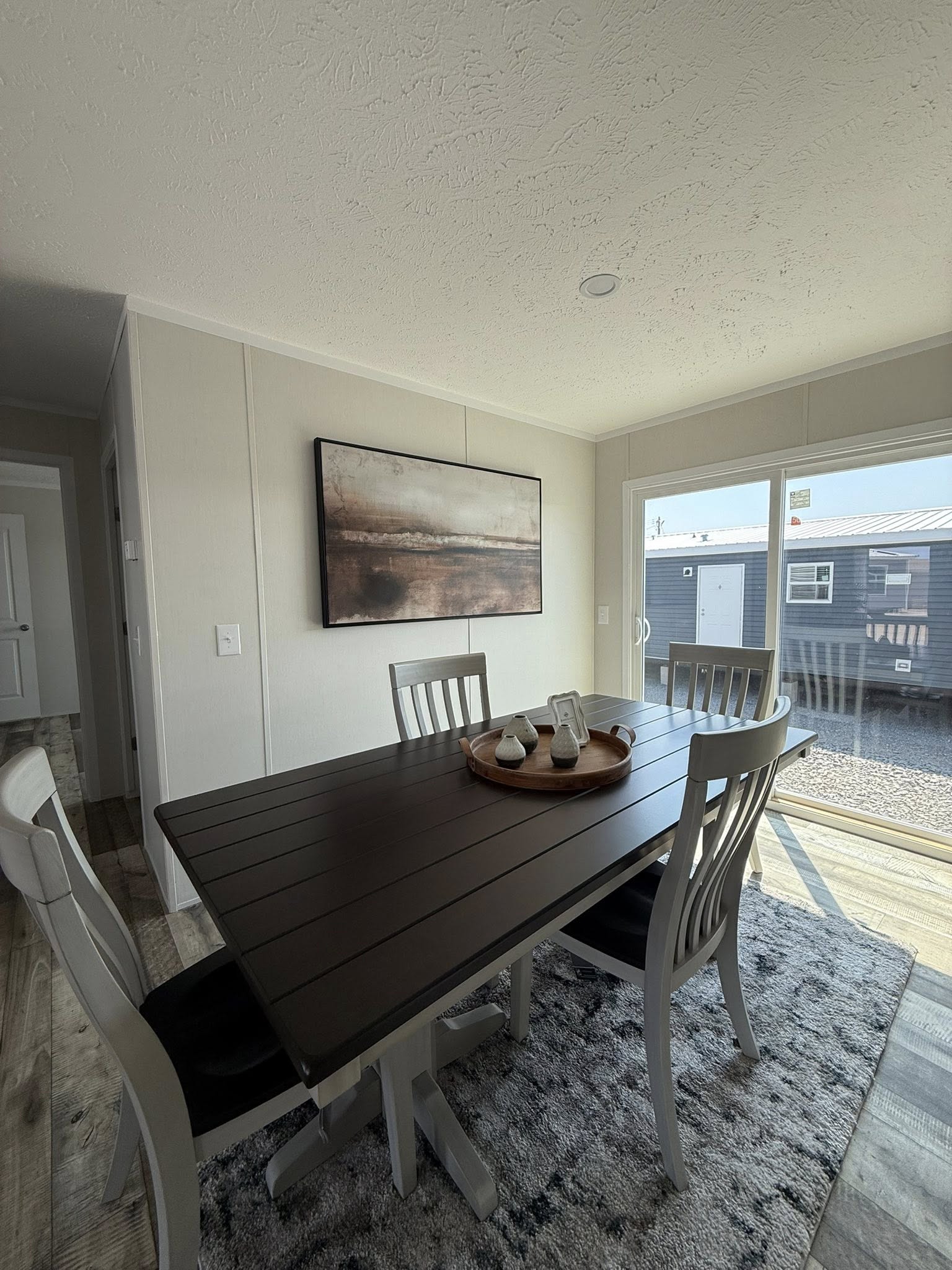 Bright dining room with a dark wooden table, four chairs, and a tray with decor. Large window letting in natural light, abstract art on the wall.