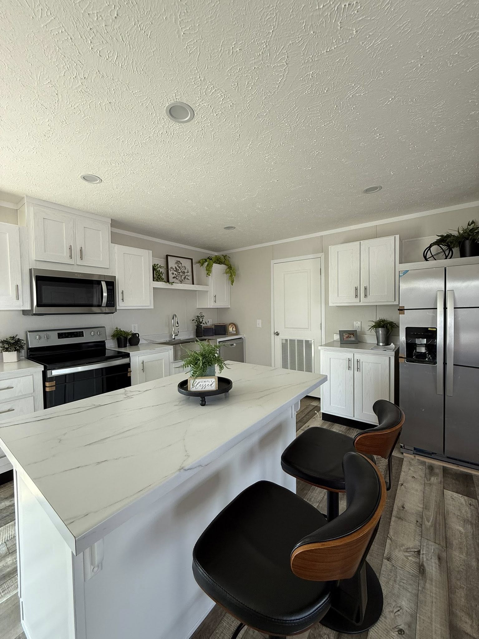 Modern kitchen with white cabinets, stainless steel appliances, and a marble island with two barstools. Potted plants add a fresh touch.