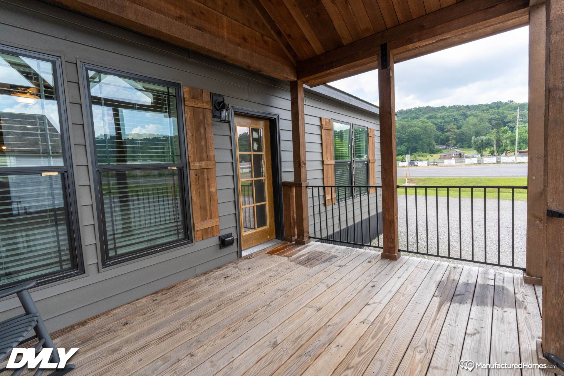Cozy wooden porch with black railing, large windows, and a glass door. Overlooks a grassy area and road, surrounded by trees under a cloudy sky.