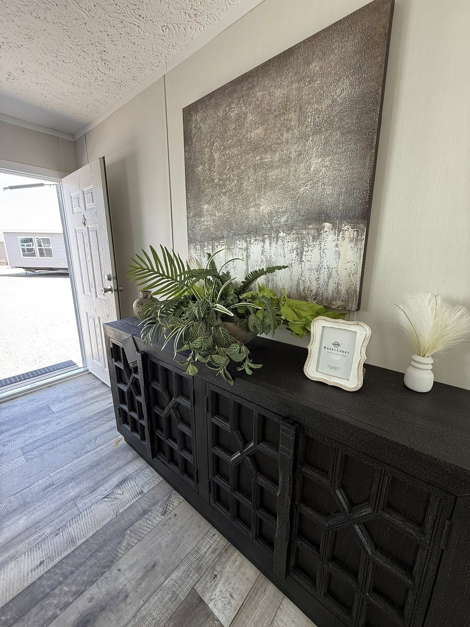 Entryway with a dark geometric cabinet holding a lush green plant and a small framed note. Neutral tones with an abstract painting above convey a modern, welcoming vibe.
