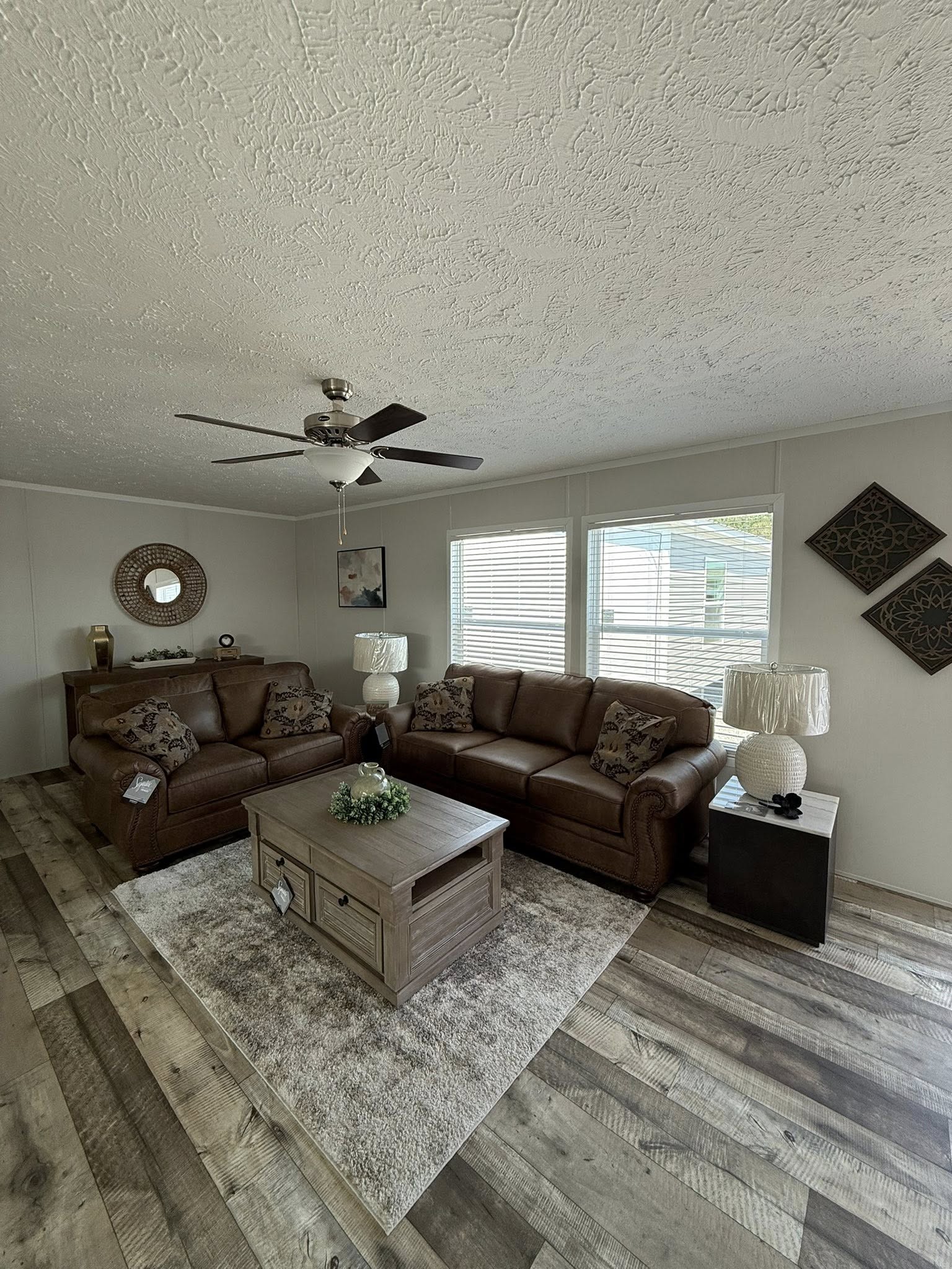 Cozy living room with two brown leather sofas, a wooden coffee table, and a plush rug on wooden flooring. Sunlit with decor on walls and soft lighting.