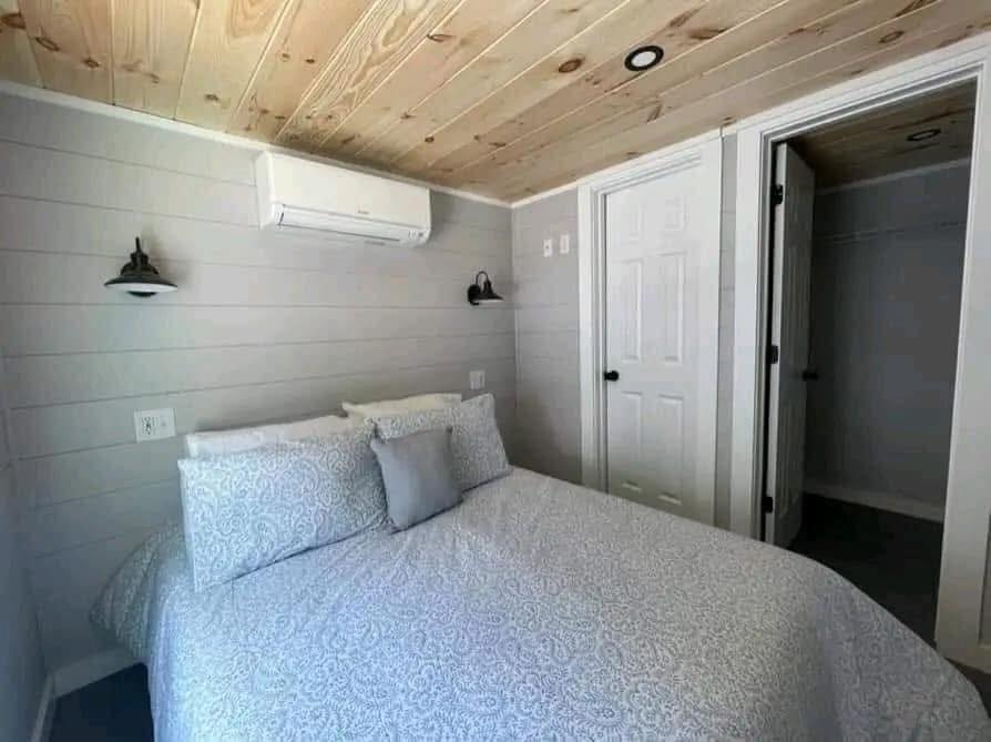 Cozy bedroom with a patterned bedspread and pillows, wood-paneled ceiling, and modern white and gray tones. Air conditioner above, doorways to the right.