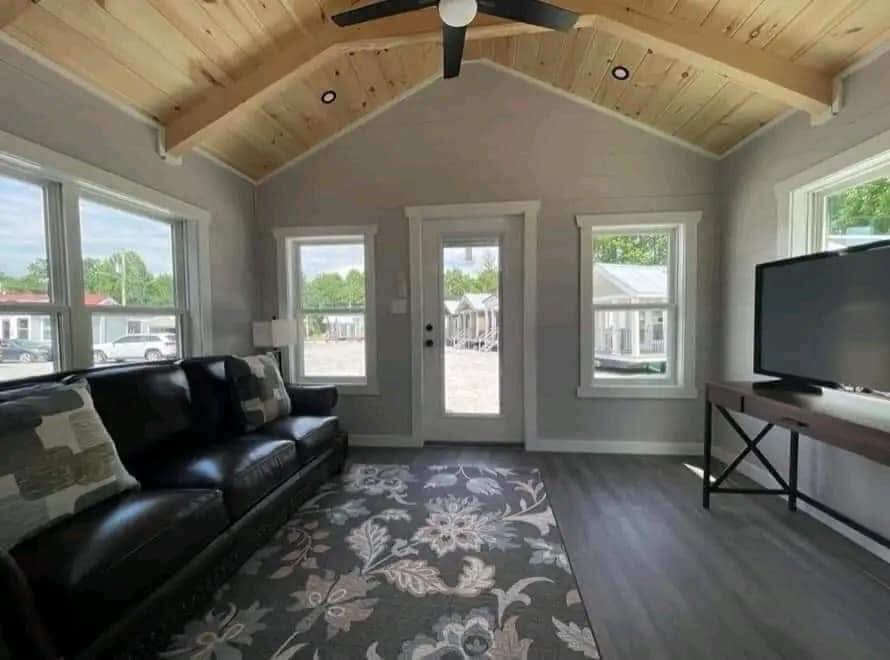 Bright living room with vaulted wooden ceiling, large windows, a dark leather sofa, floral-patterned rug, and a flat-screen TV on a stand. Cozy and modern.