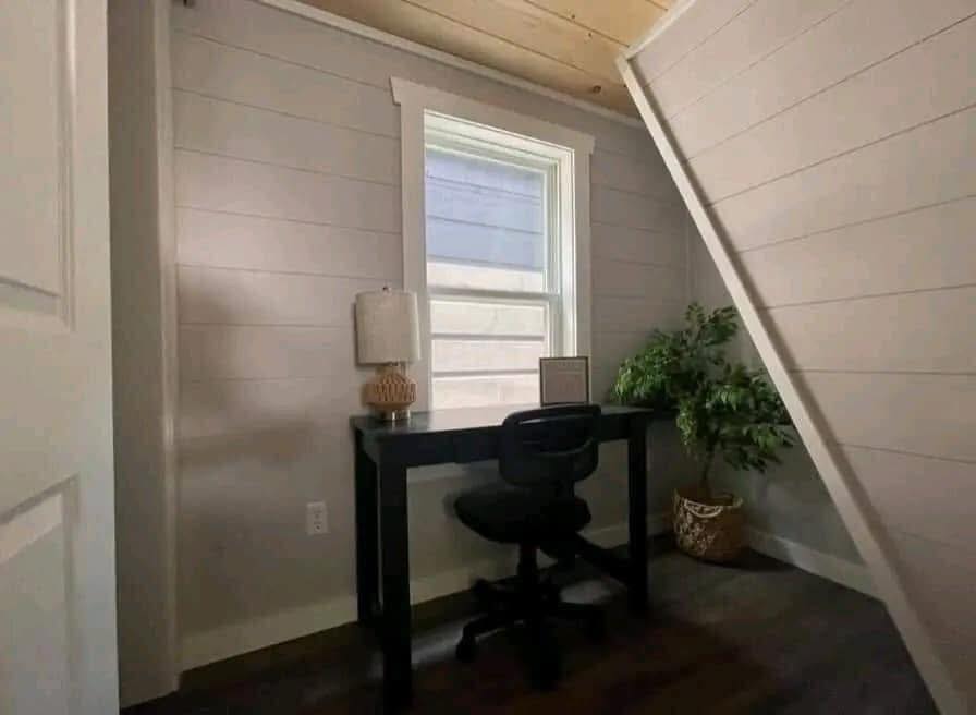 Small home office with a black desk under a slanted ceiling. A chair, lamp, and plant add coziness. Sunlight filters through the window, creating warmth.
