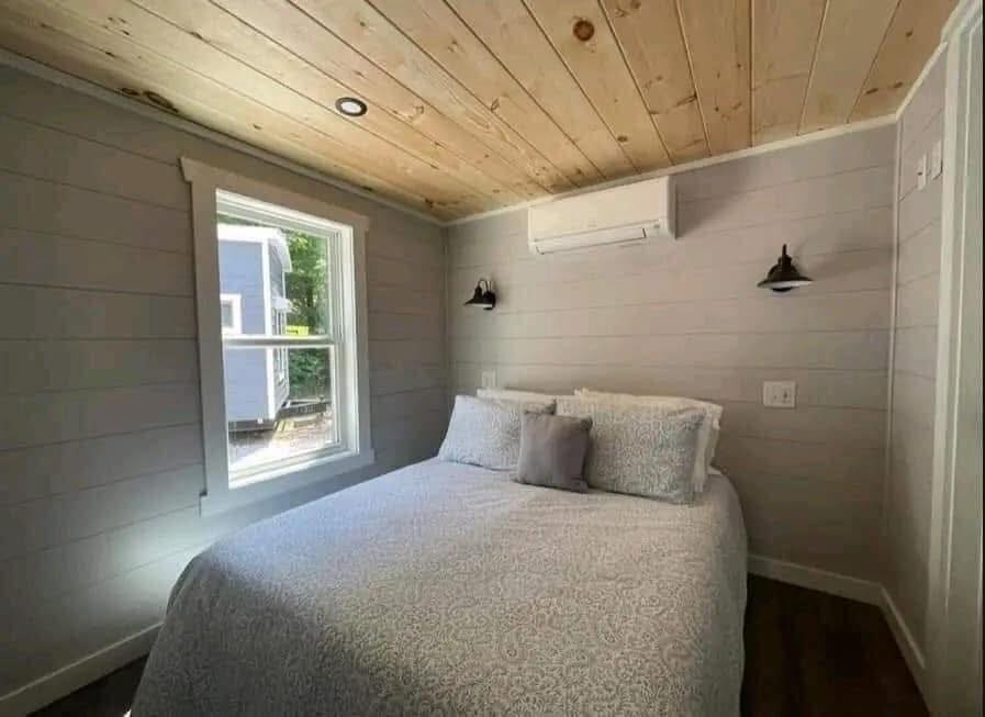 Cozy bedroom with wooden ceiling, light gray walls, and wood floors. A bed with patterned bedding faces a window. Soft lighting conveys a peaceful tone.