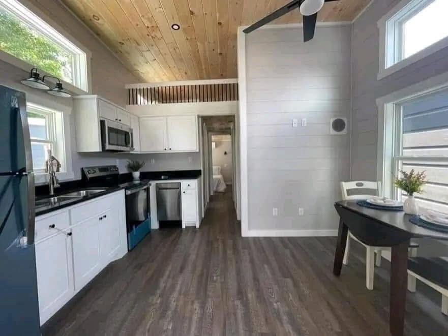 Modern tiny home interior with wood ceiling, white kitchen cabinets, stainless steel appliances, and small dining area. Bright, minimalist, cozy.