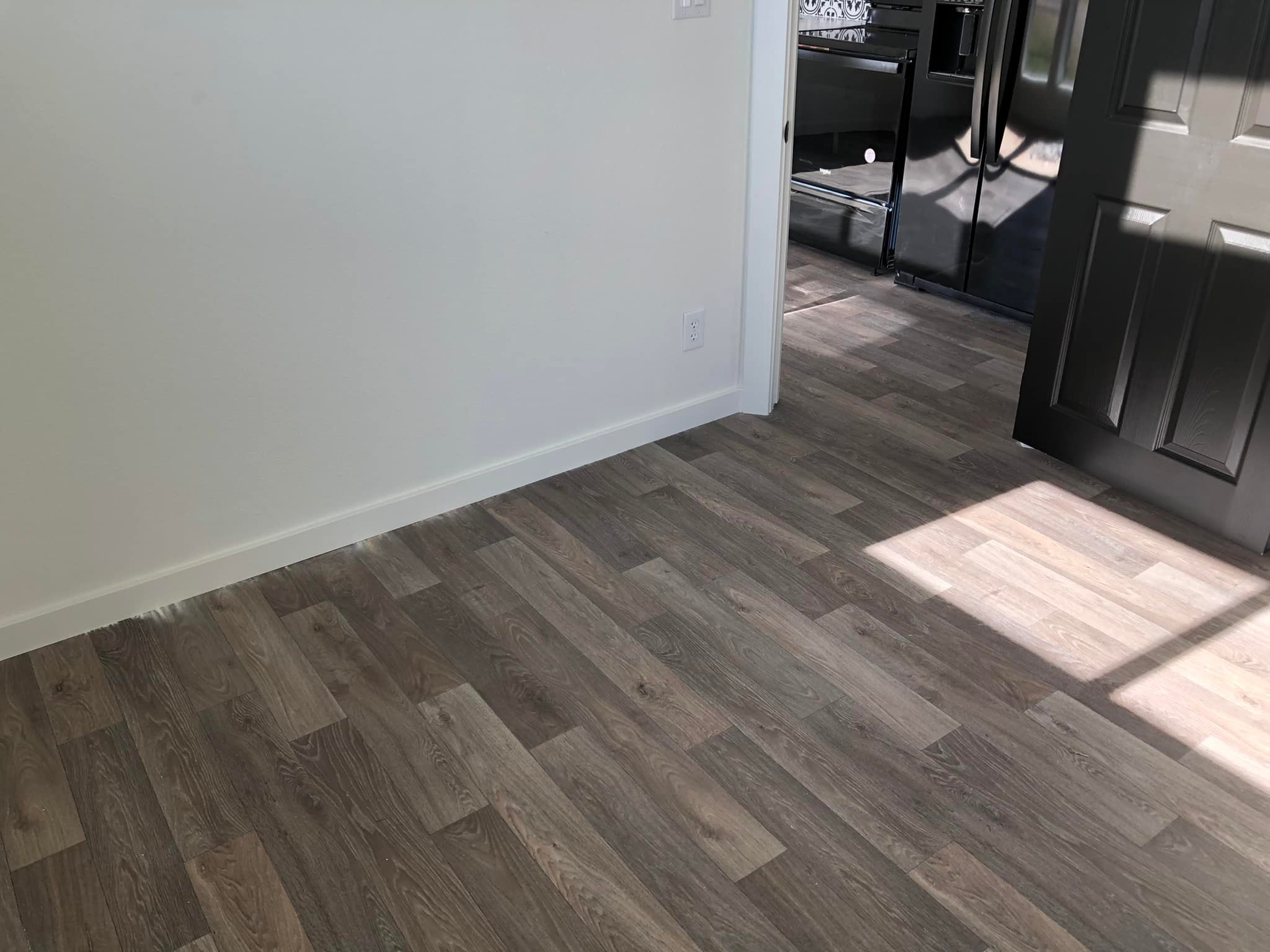 Sunlight streams onto a room with light brown wood flooring, white walls, and an open black door. A glimpse of a modern kitchen is visible.