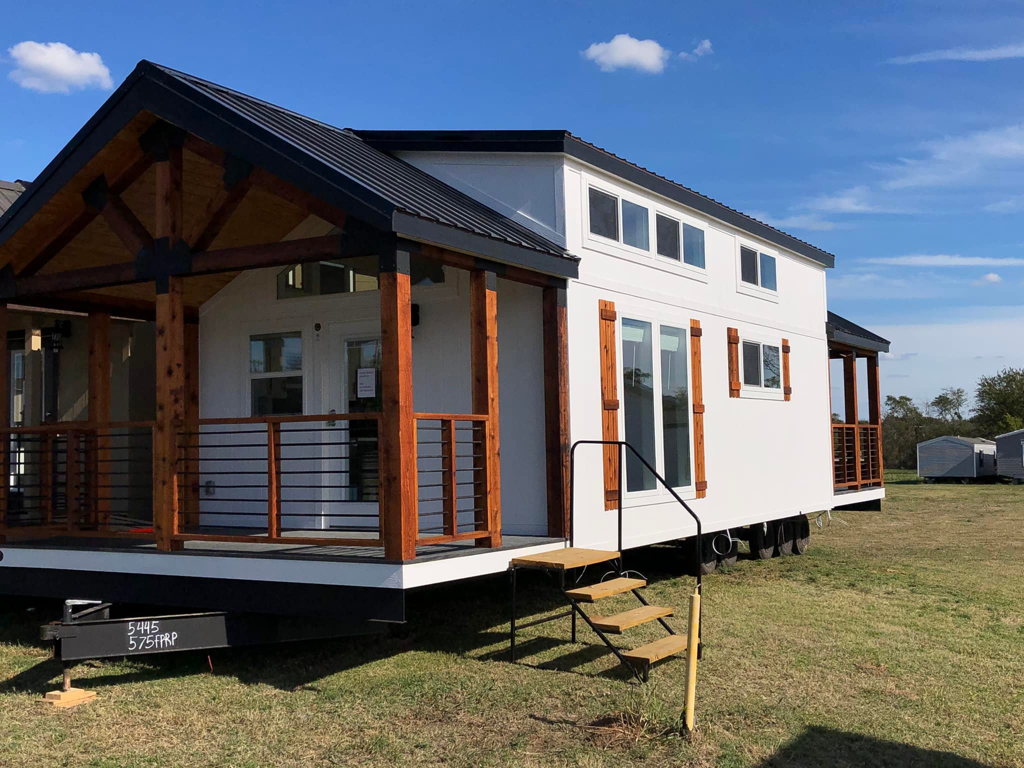 Modern tiny house with white siding, wooden accents, and a black metal roof. The porch is framed with wooden beams. Set on grass under a clear blue sky.