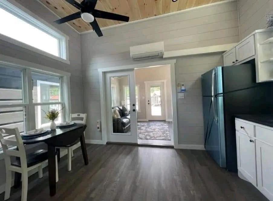 Bright kitchen with a wooden ceiling and fan, featuring a dining table by a window, white cabinets, a black fridge, and glass doors leading to a cozy living room.