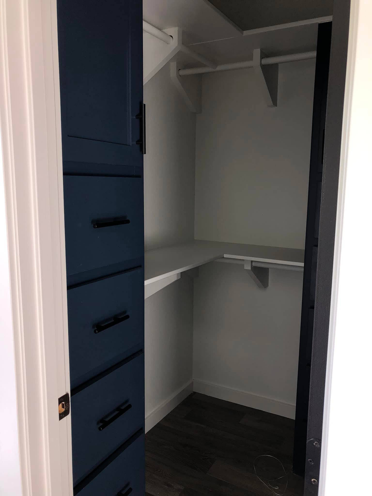 Small walk-in closet with blue cabinets and dark handles on the left. White shelves and hanging rods extend along the back and right walls. Dark wood floor.