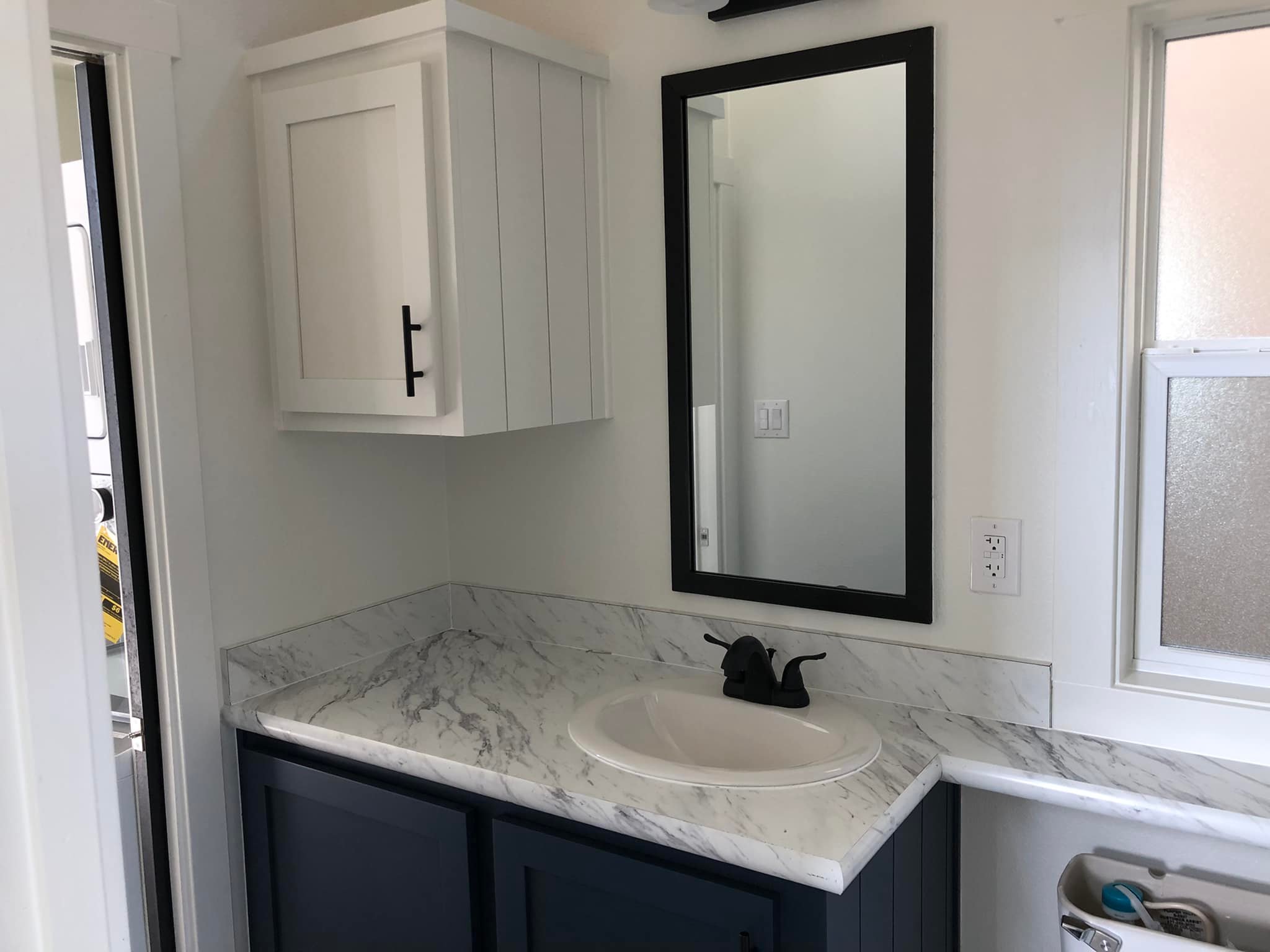 Compact bathroom with white and navy color scheme, featuring a marble countertop, white cabinet, black-framed mirror, and window, creating a sleek, modern vibe.