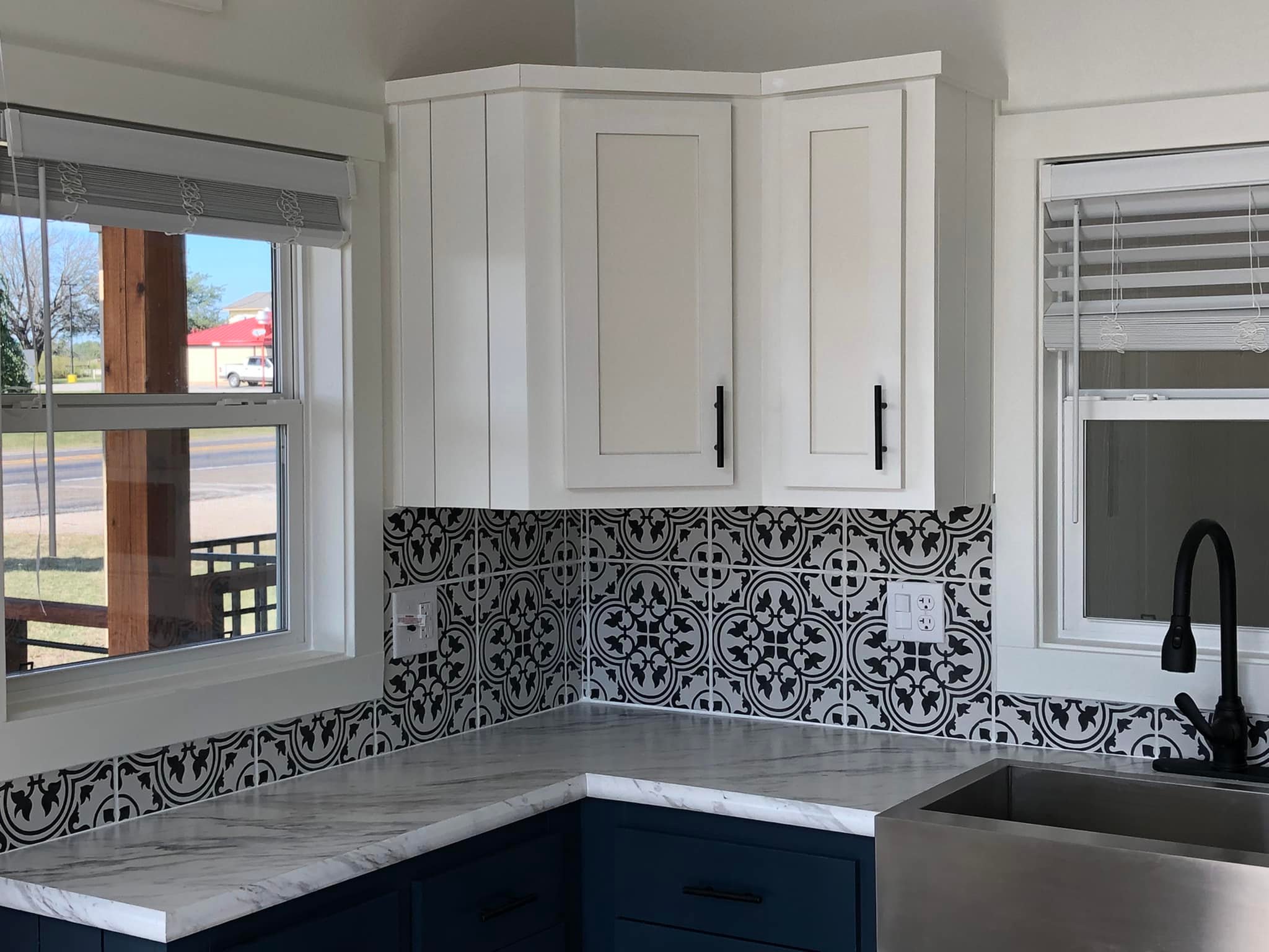 A modern kitchen corner with white upper cabinets and dark handles, blue lower cabinets, patterned black and white backsplash, and a marble countertop. Two windows offer a bright, airy feel.