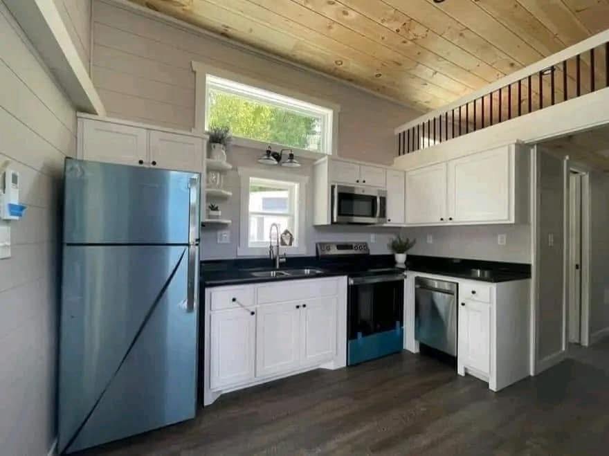 Compact kitchen with white cabinets, black countertops, a stainless steel fridge, microwave, and dishwasher. Light wood ceiling, warm and cozy feel.