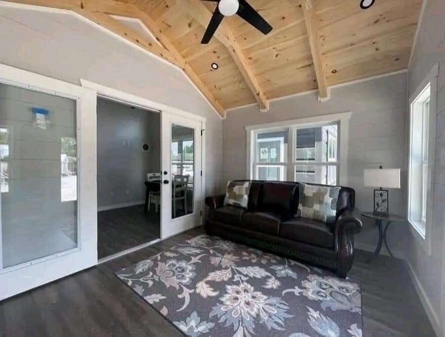 Cozy living room with wooden vaulted ceiling, brown leather sofa adorned with patterned cushions, floral rug, and a lamp on a side table. Bright and airy ambiance.