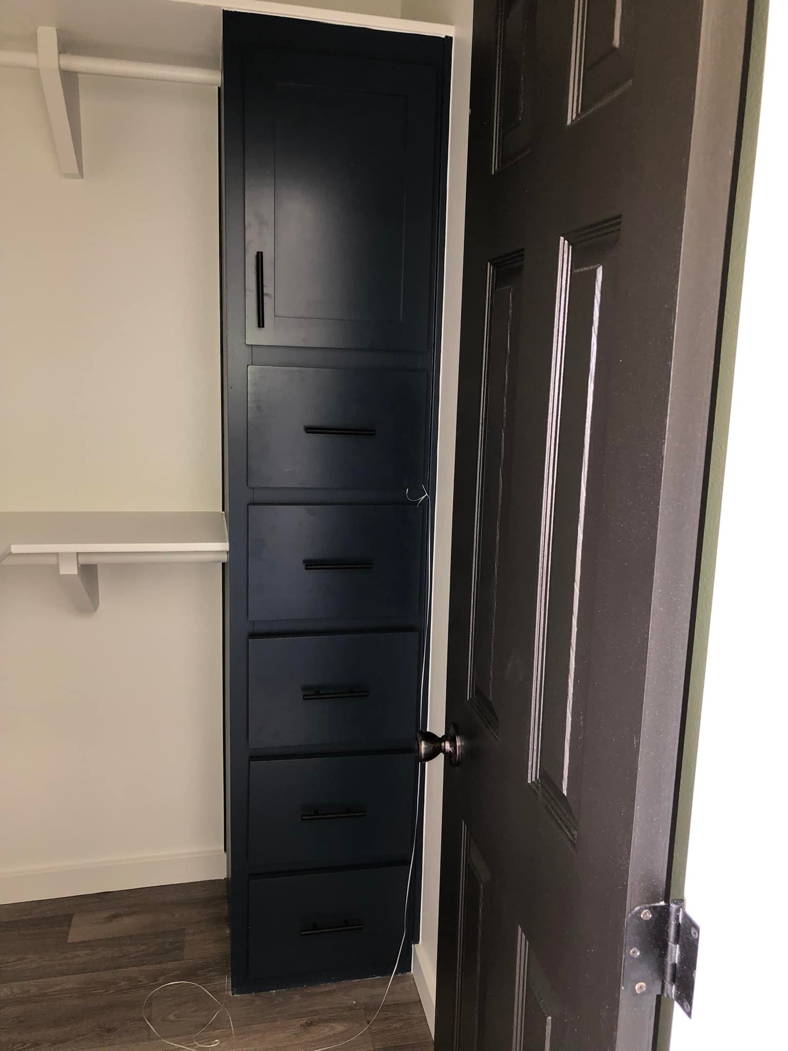 Narrow closet with dark navy cabinets and black handles, partially hidden by an open dark brown door. White shelves are visible beside the cabinet.