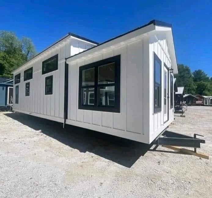 A white tiny house with black trim features large windows and a gabled roof, set on a gravel lot under a clear blue sky, surrounded by trees.