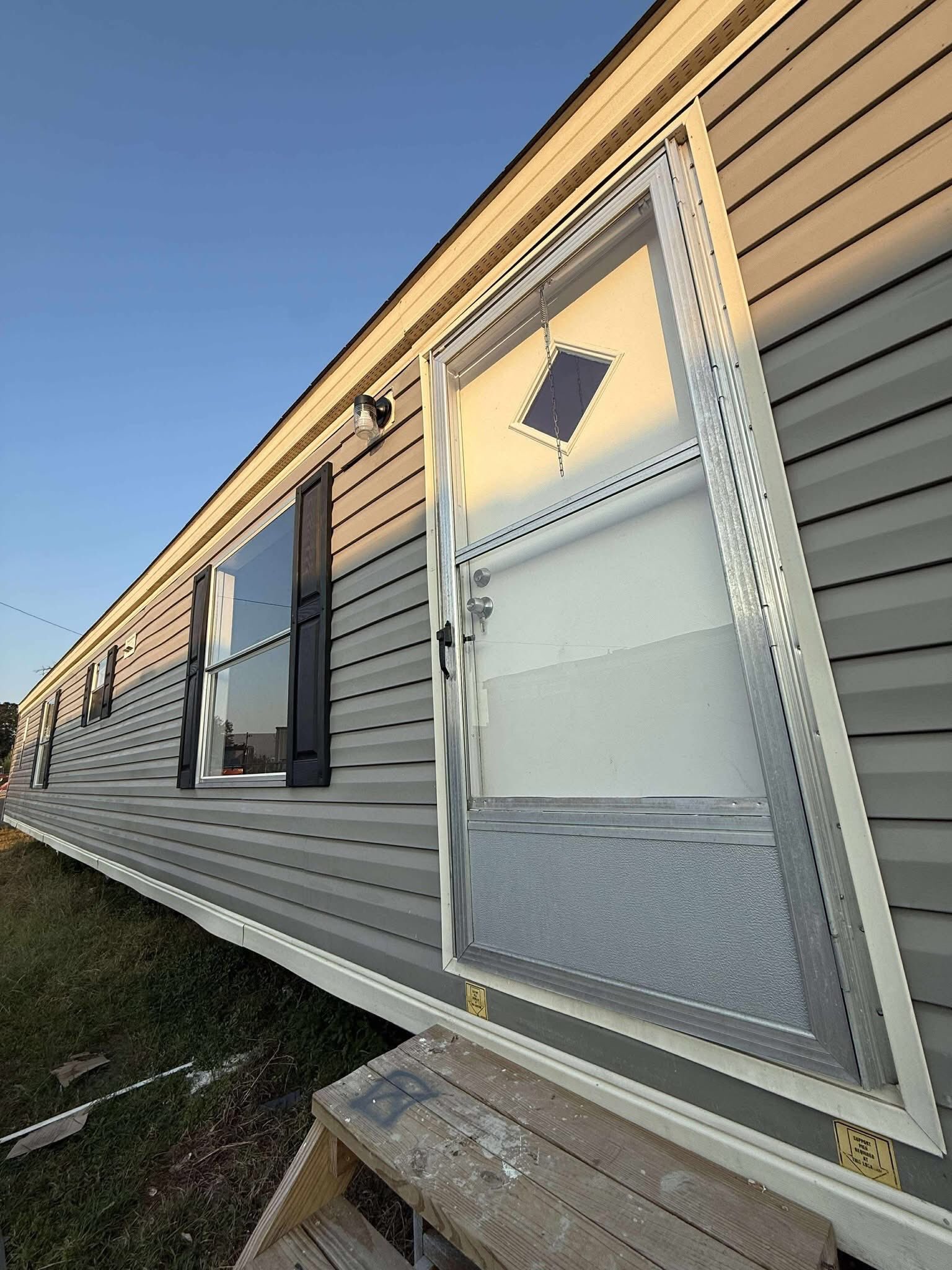 Tilted view of a gray mobile home with a door and two windows. The door is misaligned, creating a surreal effect. Early evening light casts shadows.