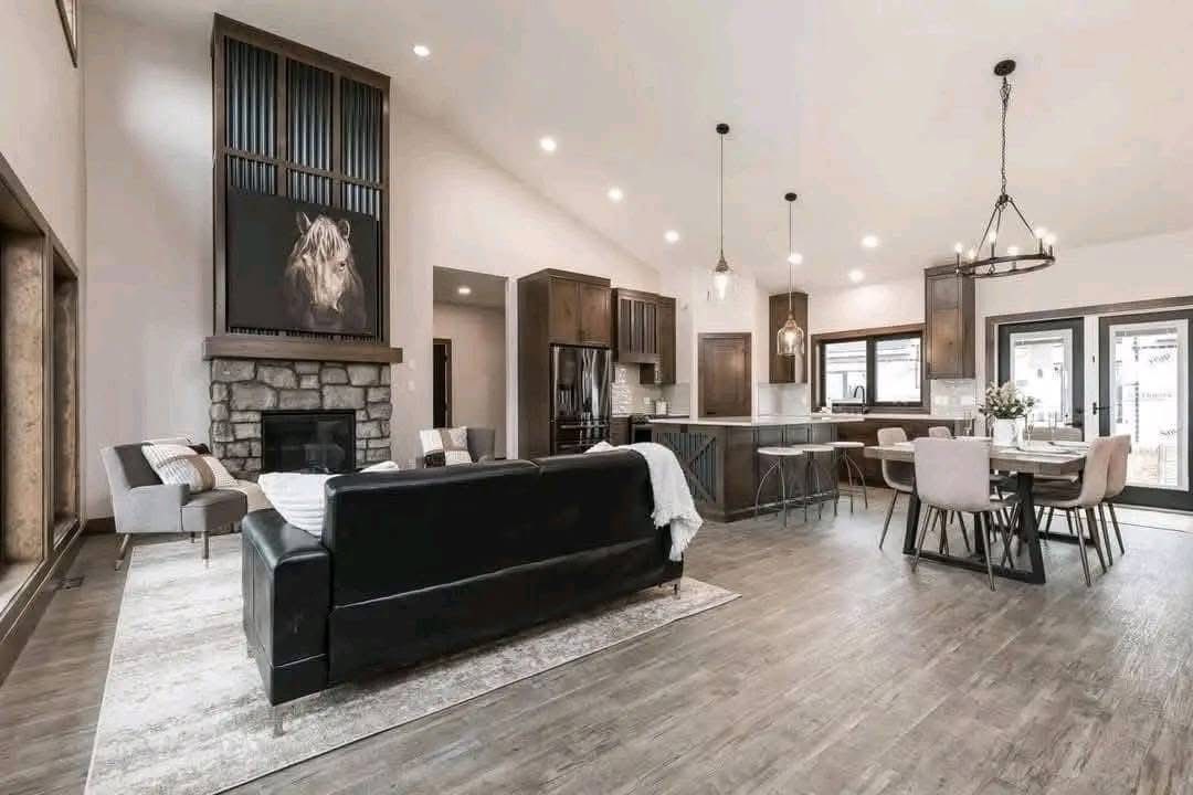 Spacious open-plan living room and kitchen with high ceilings. Features a modern design, stone fireplace, cozy seating, and a dining area under a chandelier.