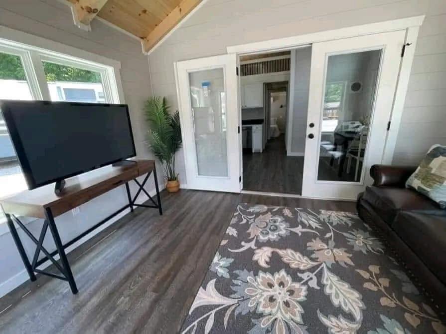 A cozy living room with a dark wood floor, patterned rug, and a brown leather couch. A flat-screen TV is on a wooden stand. Glass doors lead to a kitchen.
